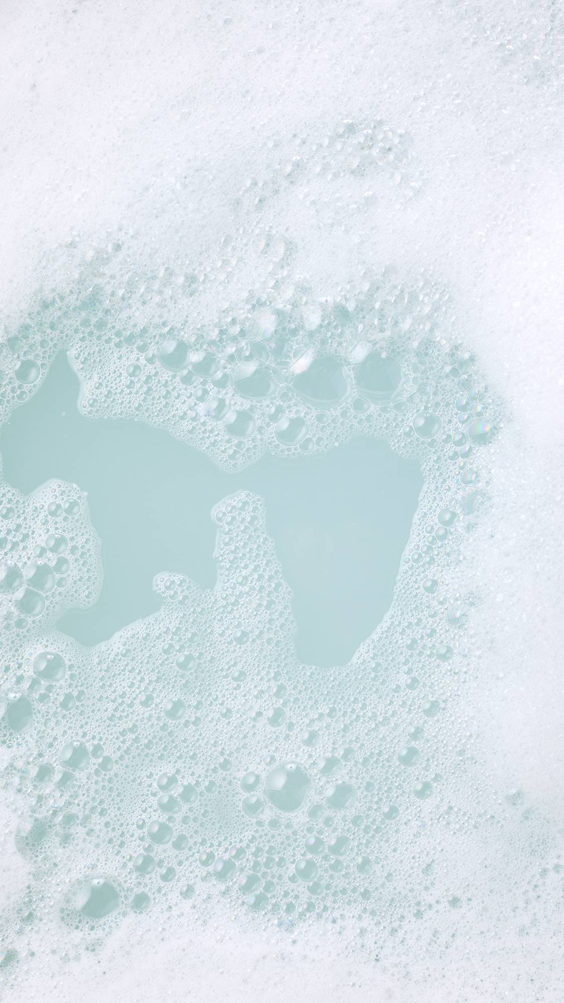 A dense layer of soft white bath foam parts across clear, pale duck-egg blue water, surrounded by clusters of fine, iridescent bubbles.
