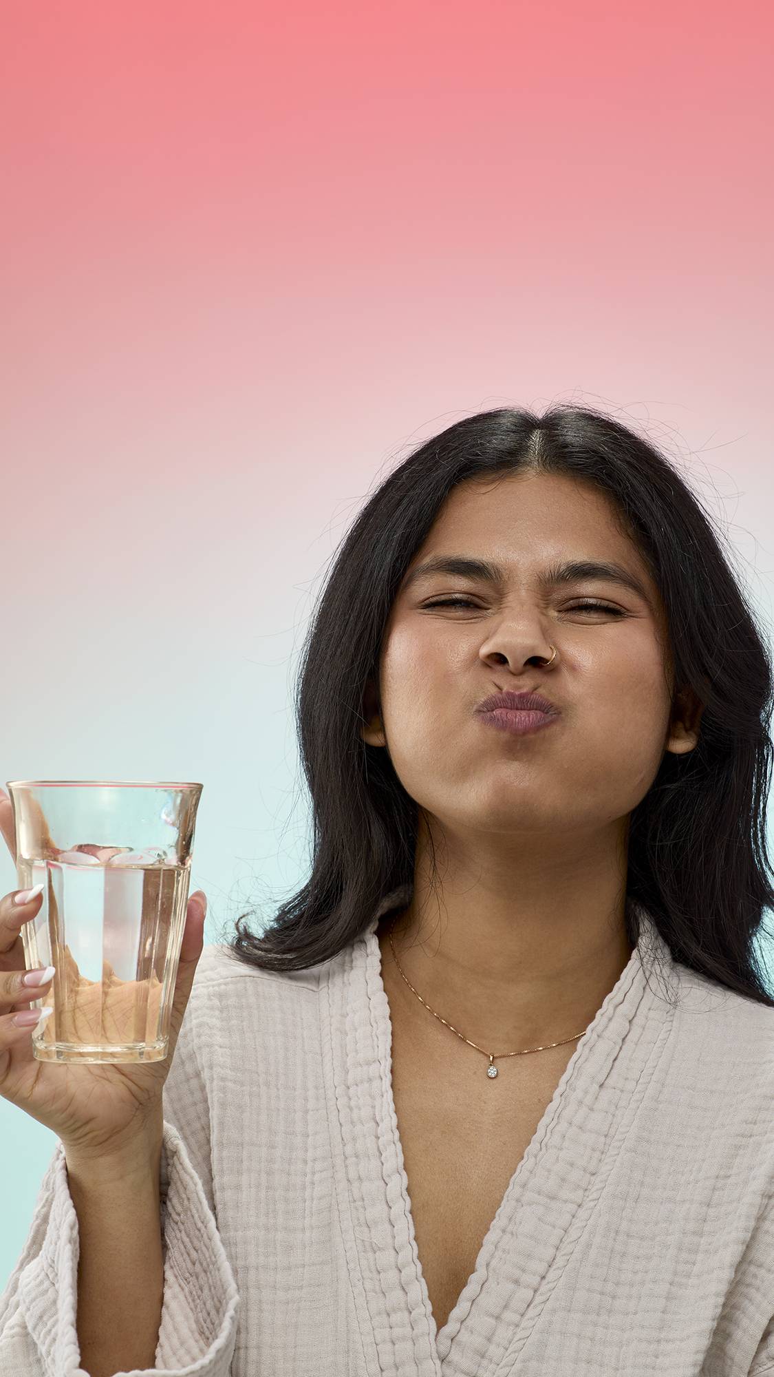 A person with long dark hair swishes mouthwash with puffed-out cheeks and squinted eyes while holding a clear glass of water.