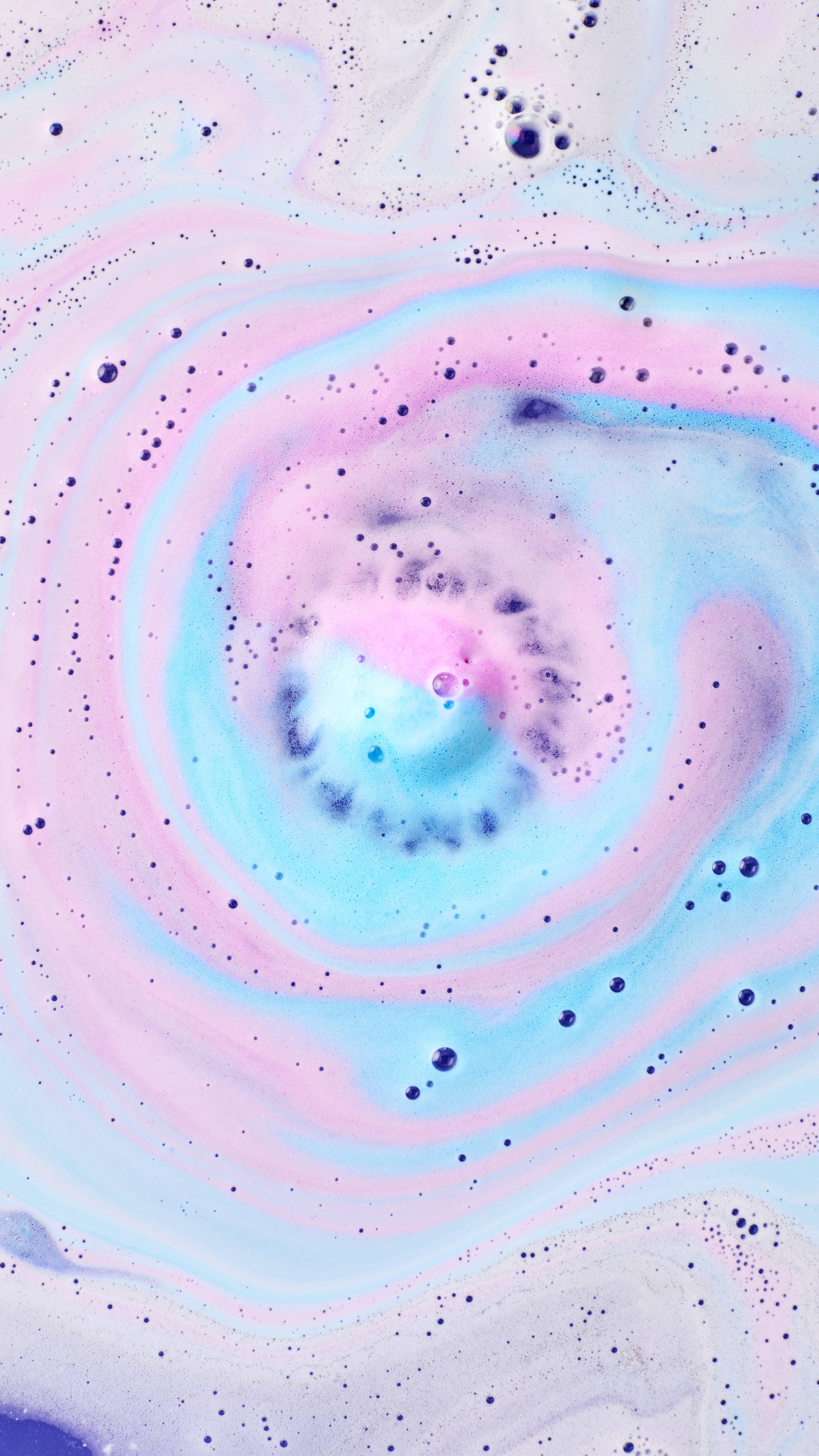 An aerial view of bathwater featuring a central swirl of pink, blue, and lilac foam, created by the "Bathe All The Time" bath bomb. Tiny bubbles and delicate frothy patterns spiral outwards, creating a dreamlike, marbled effect across the surface.