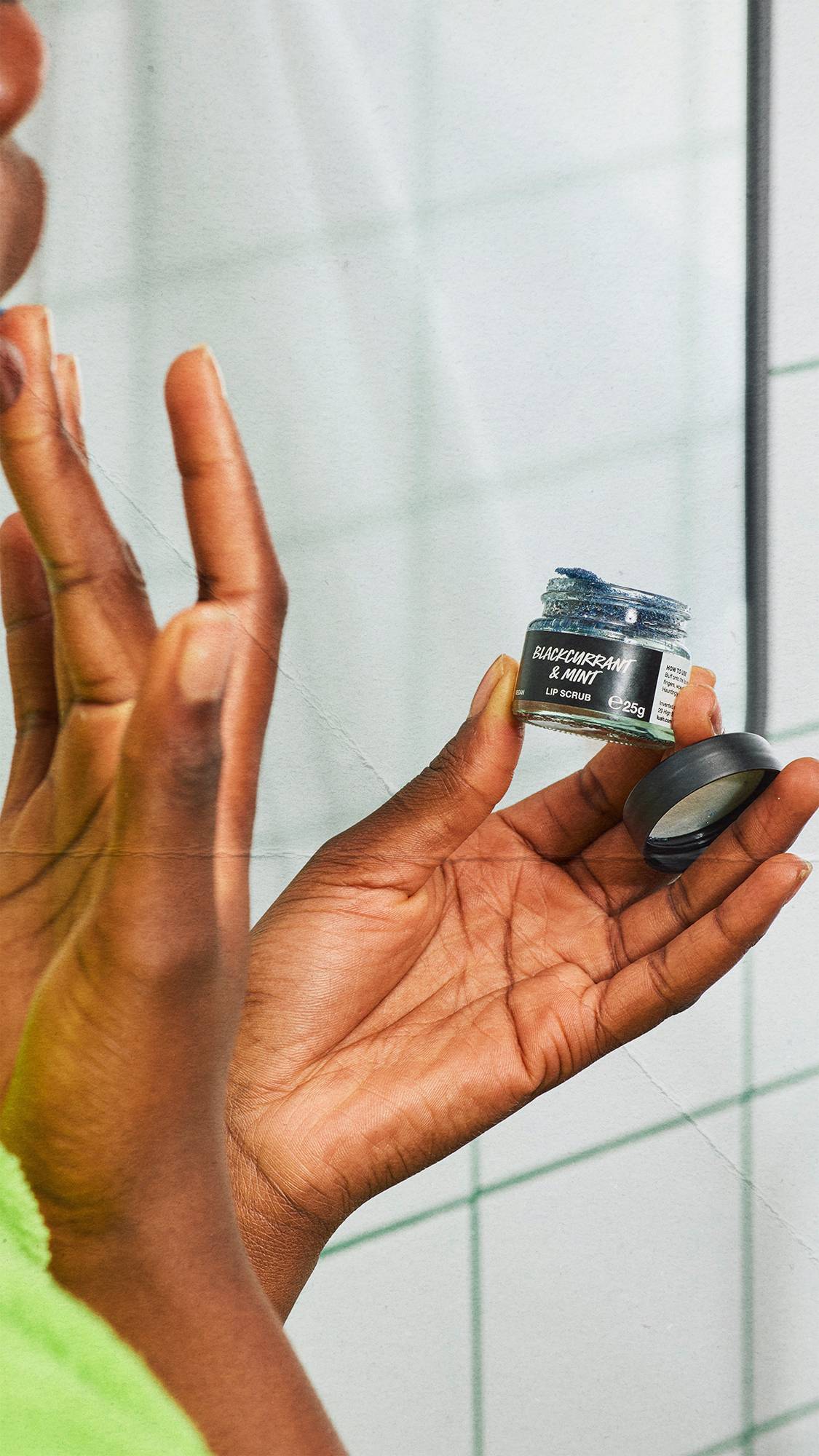 A close-up shot of a person's hands holding an open small glass pot of Lush 'BLACKCURRANT & MINT' lip scrub, which contains a dark, shimmering blue product. White square tiles with green grout lines form the background.