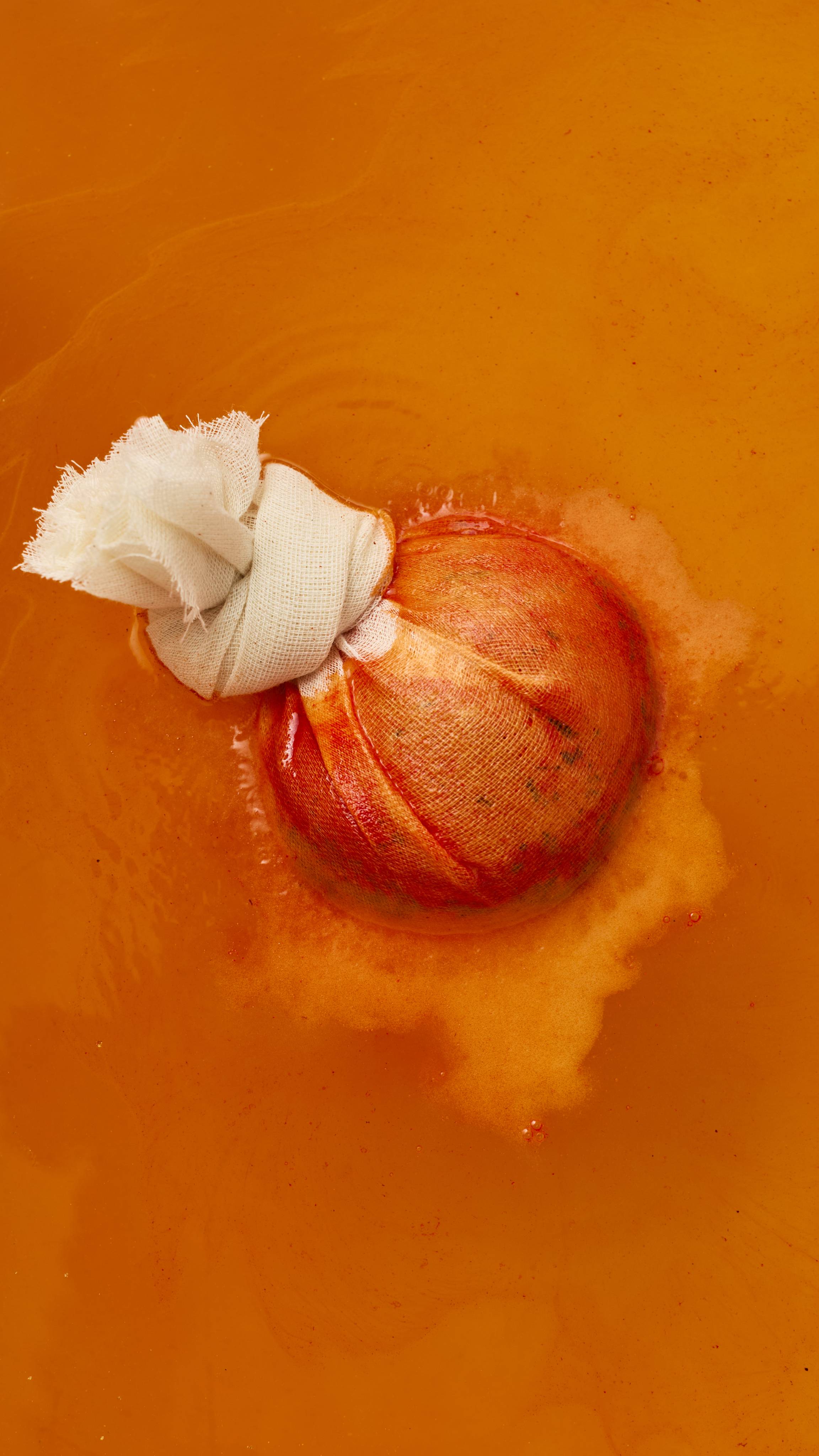 A close-up, top-down view of a round bath bomb in a tied cheesecloth pouch, actively fizzing in rich orange water. Red dish hues bleed through the fabric as a pool of orange-tinted foam surrounds the dissolving bath bomb.