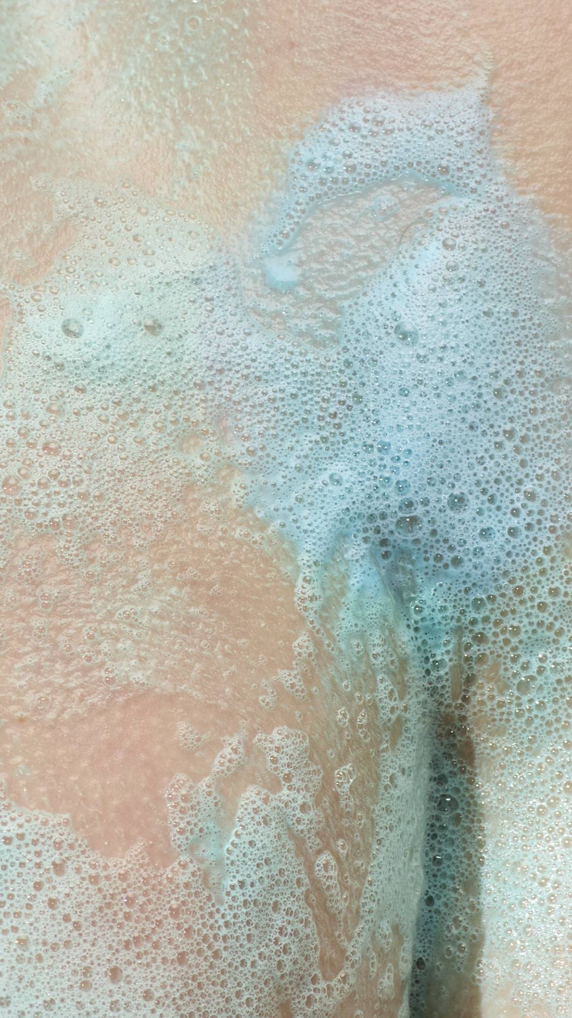 A close-up texture shot showing pale skin partially covered in a thick, bubbly layer of light blue soap lather. Fine suds and larger translucent bubbles stretch across the surface.