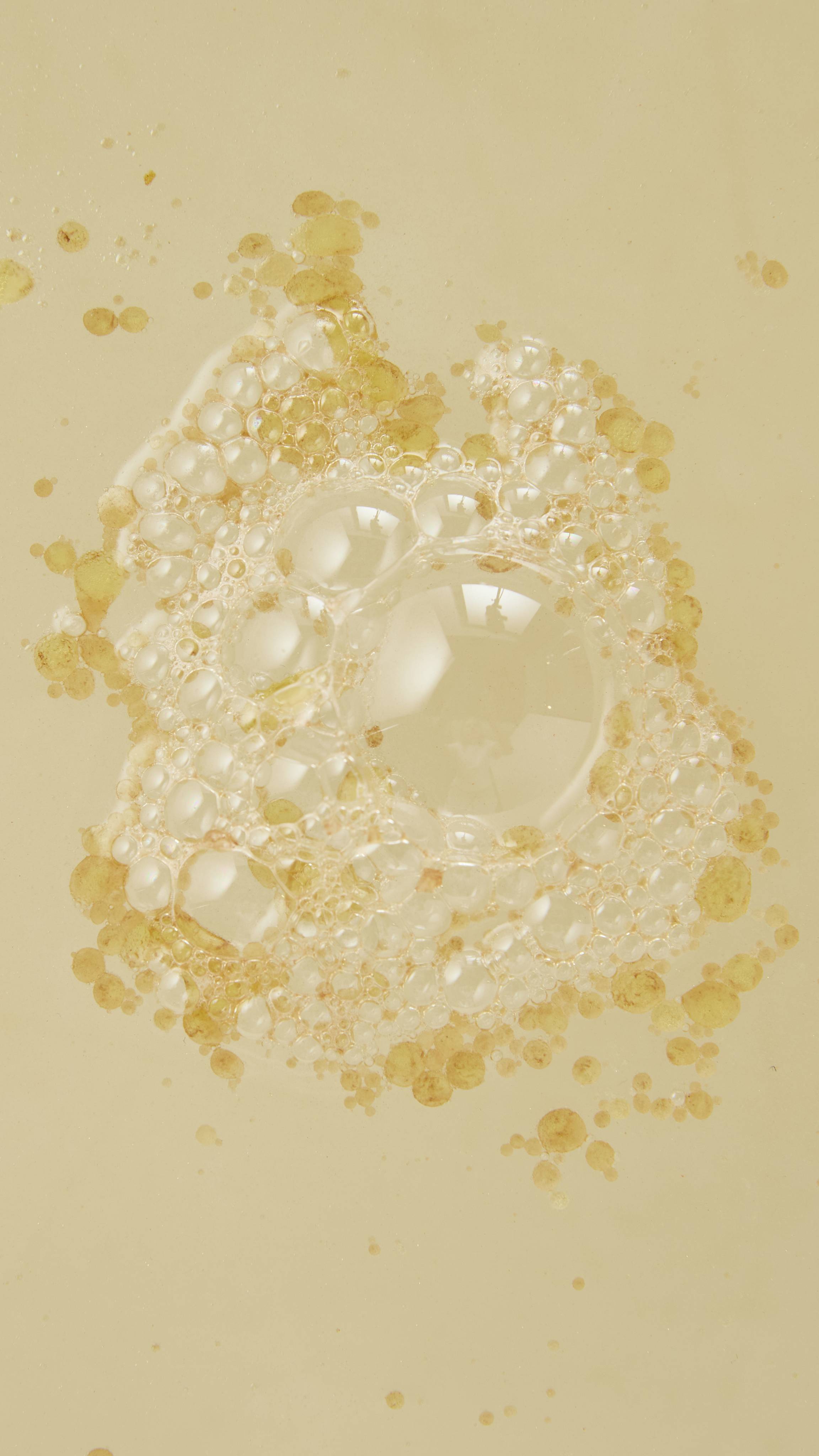 An aerial view of the bathwater after using the Lush "Butterbear" bath bomb/ A cluster of clear, glossy bubbles surrounded by tiny, golden oil droplets floats on the surface of pale bathwater.