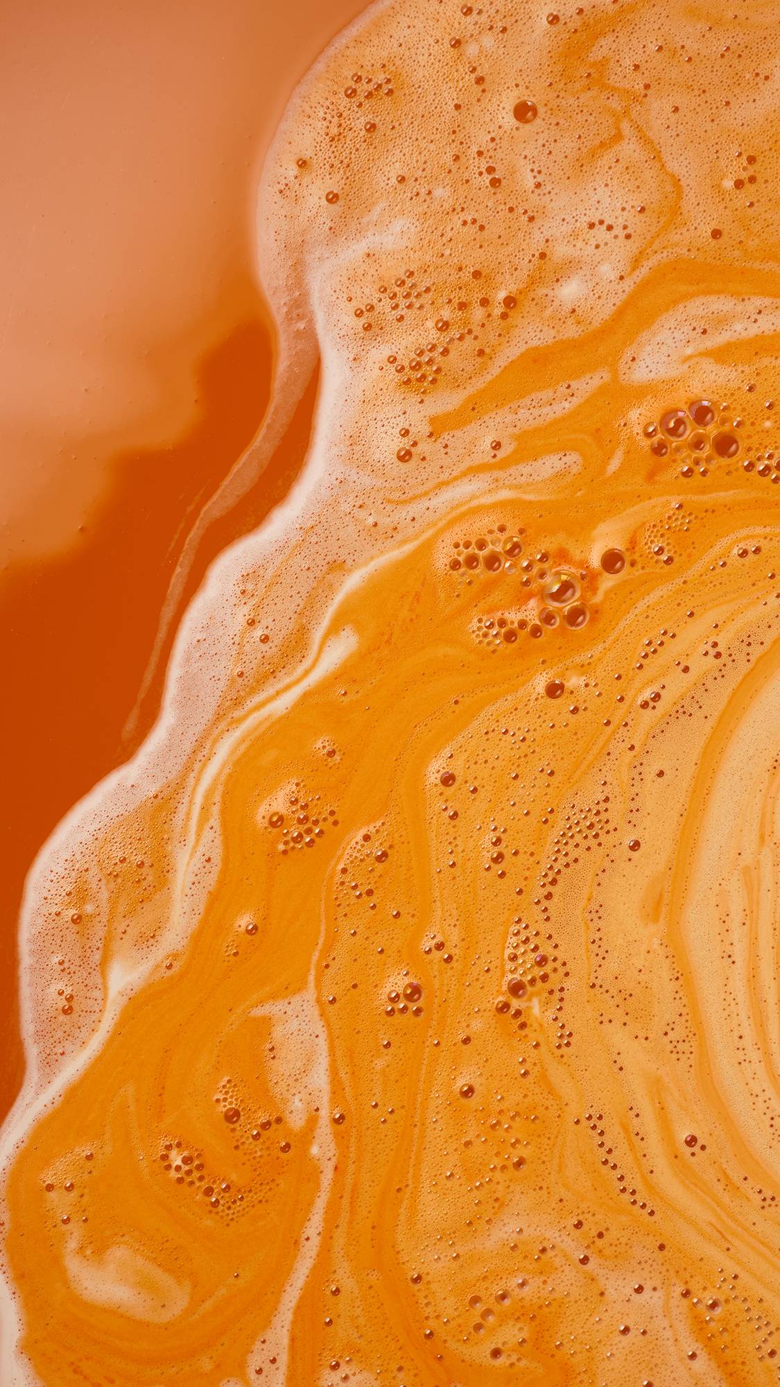 A close-up of vibrant orange bathwater featuring rich, swirling streams of creamy white foam and delicate clusters of fine bubbles across the surface.