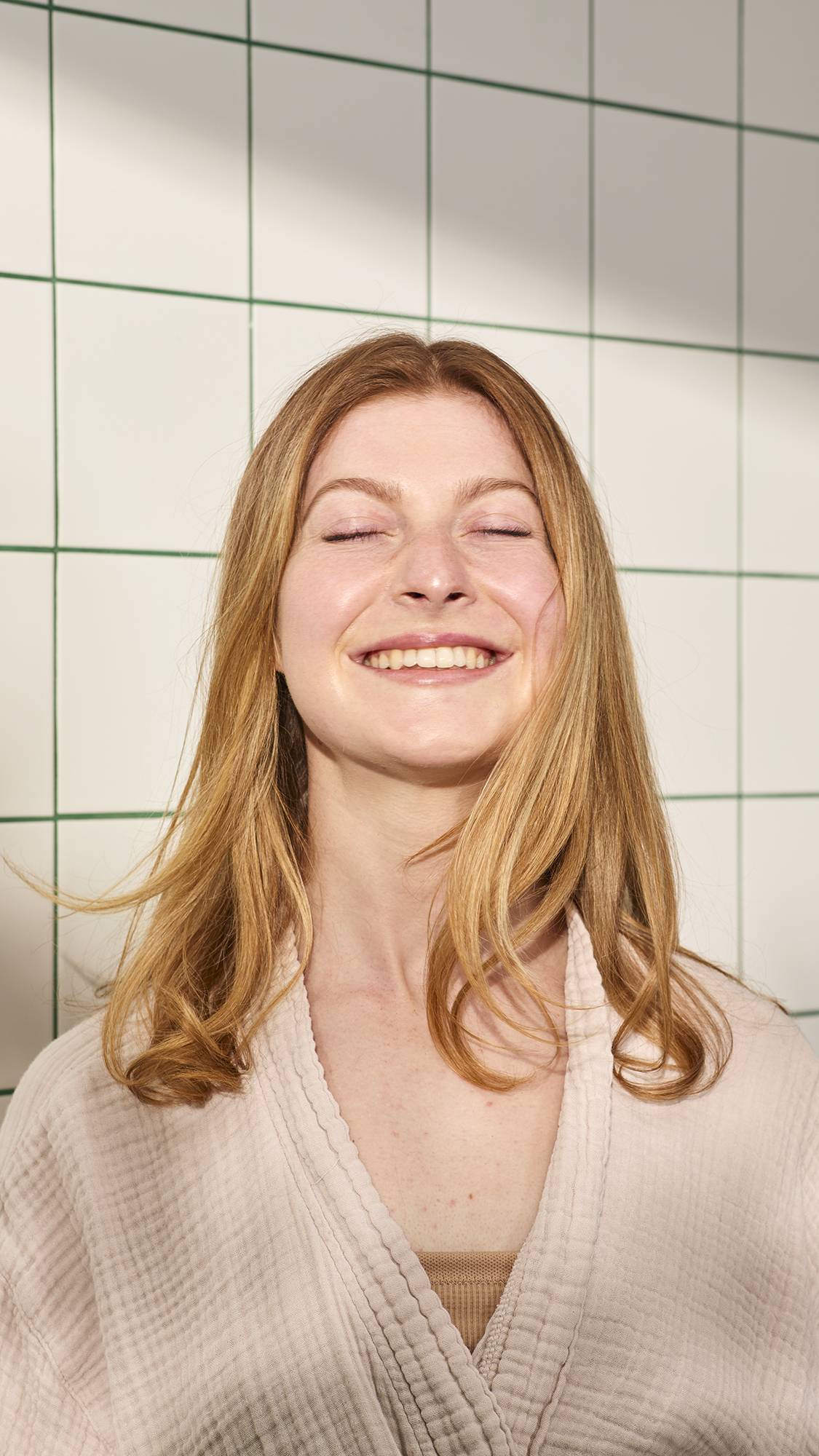 A person with medium-length , shiny, strawberry blonde hair smiles broadly with their eyes closed, wearing a light pink robe. They stand against a background of white tiles with green grout, illuminated by bright, natural light.
