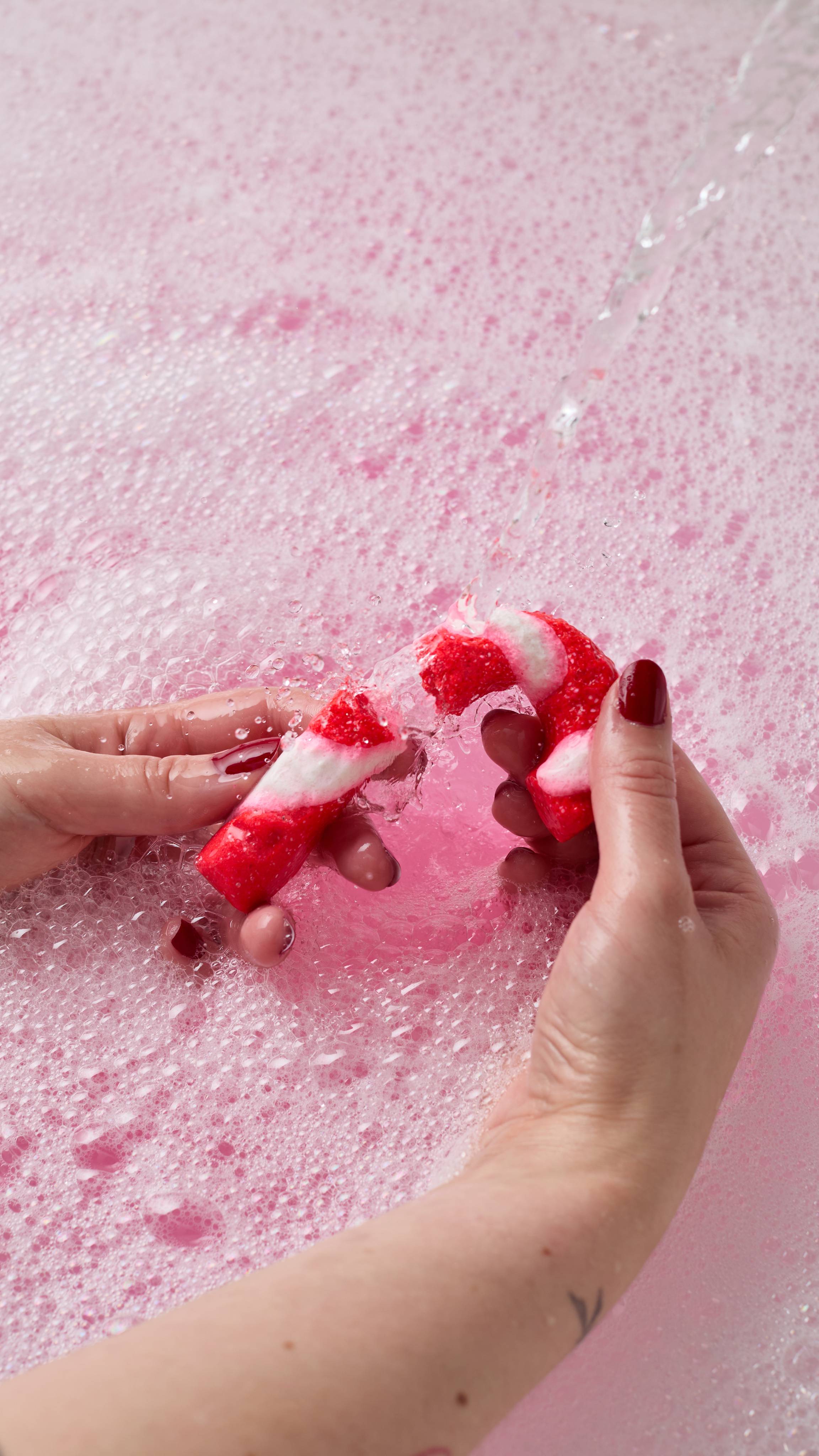 Two hands snap a red and white striped candy cane bubble bar in half under a stream of running water, surrounded by dense pink and white bath foam.