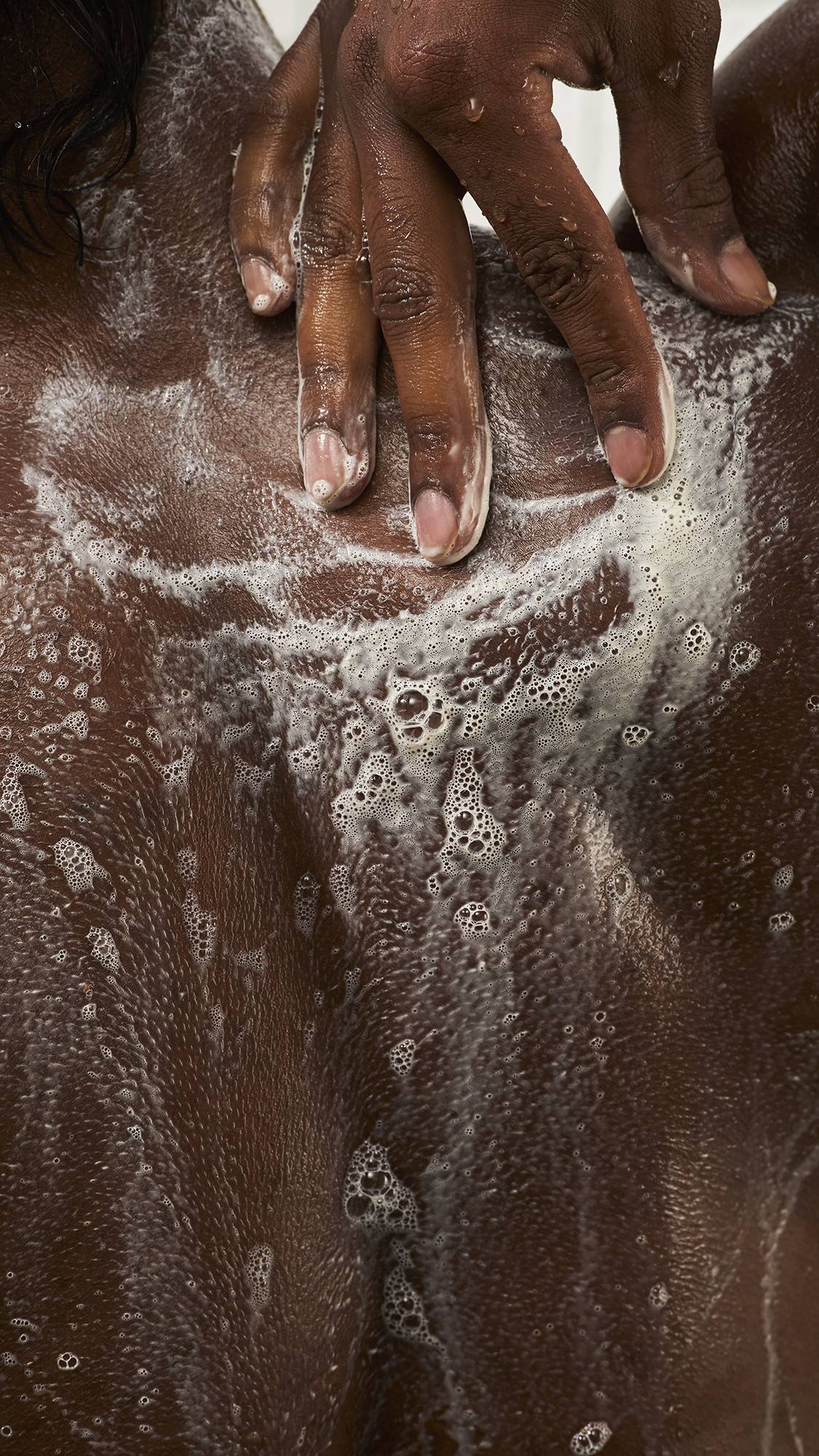 Close-up of a person smoothing rich, bubbly white soap lather across their upper back and shoulders with their hand in the shower under streaming water.