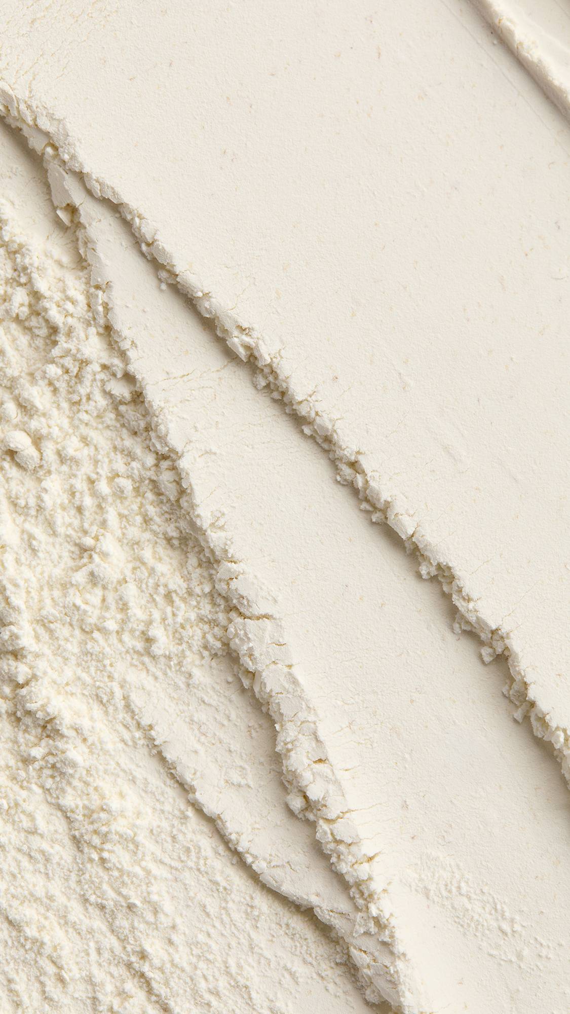 A close-up macro texture shot of finely milled, off-white "Chelsea Morning" dusting powder, displaying smooth diagonal waves alongside textured, crumbled edges.