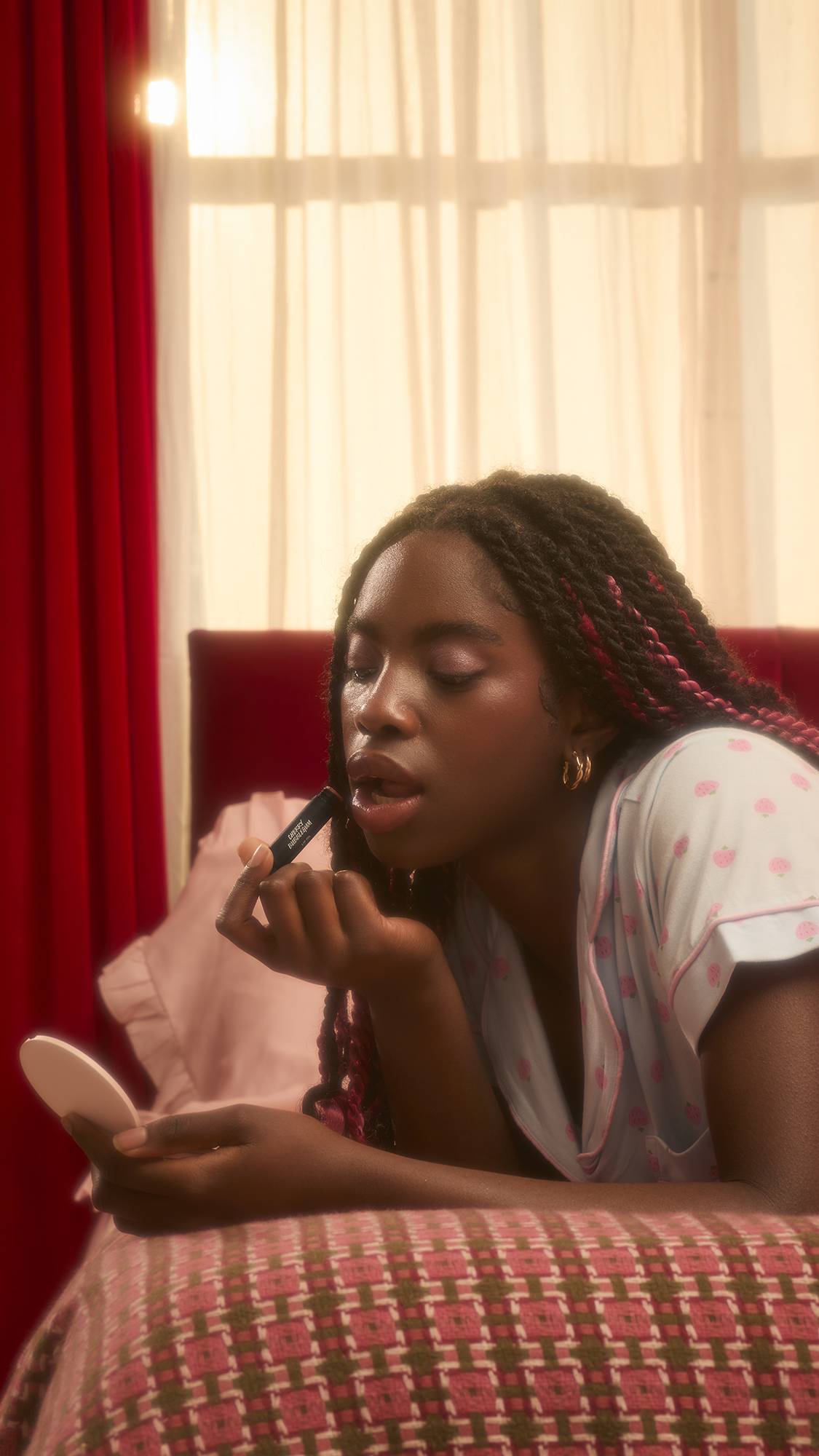 A person with long, dark braids lies on a patterned bedspread, looking into a small pink hand mirror while applying a black tube of Cherry Bubblegum solid lip oil. They are wearing light blue pyjamas with a pink strawberry pattern.