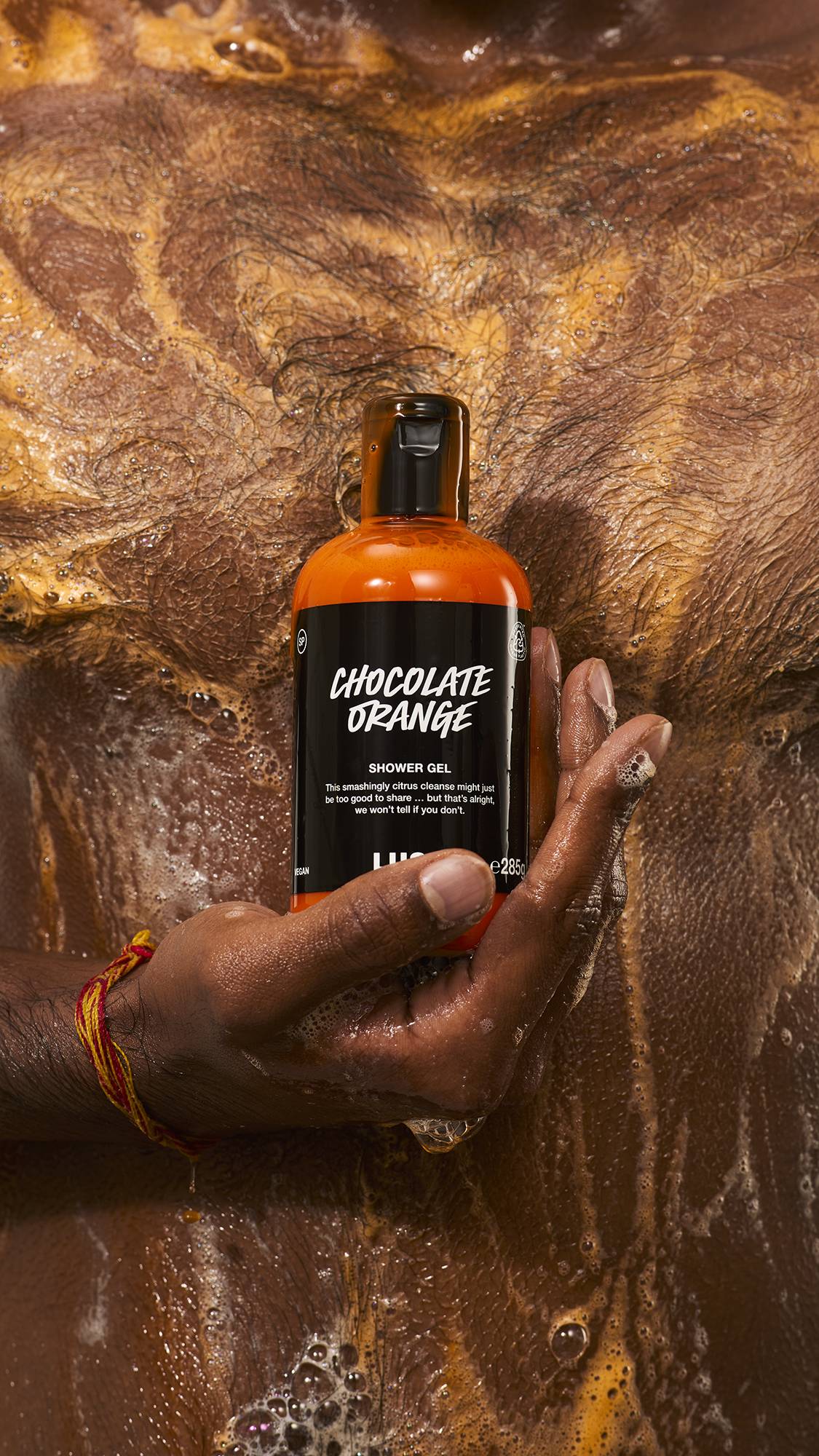 A person with dark skin holds a bottle of Lush "Chocolate Orange" shower gel against their lathered, wet chest, surrounded by rich brown and orange swirling suds.