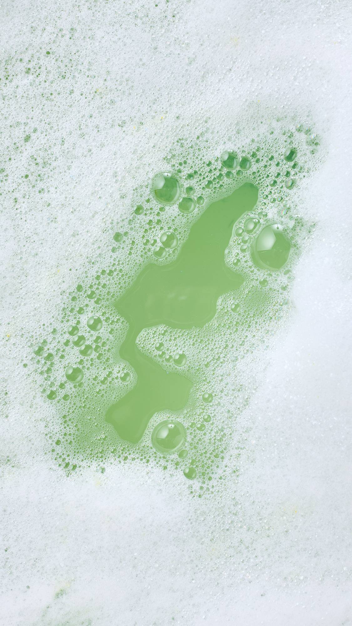 A thick blanket of creamy white bath foam parts to reveal soft green bathwater beneath, surrounded by clusters of fine frothy bubbles.