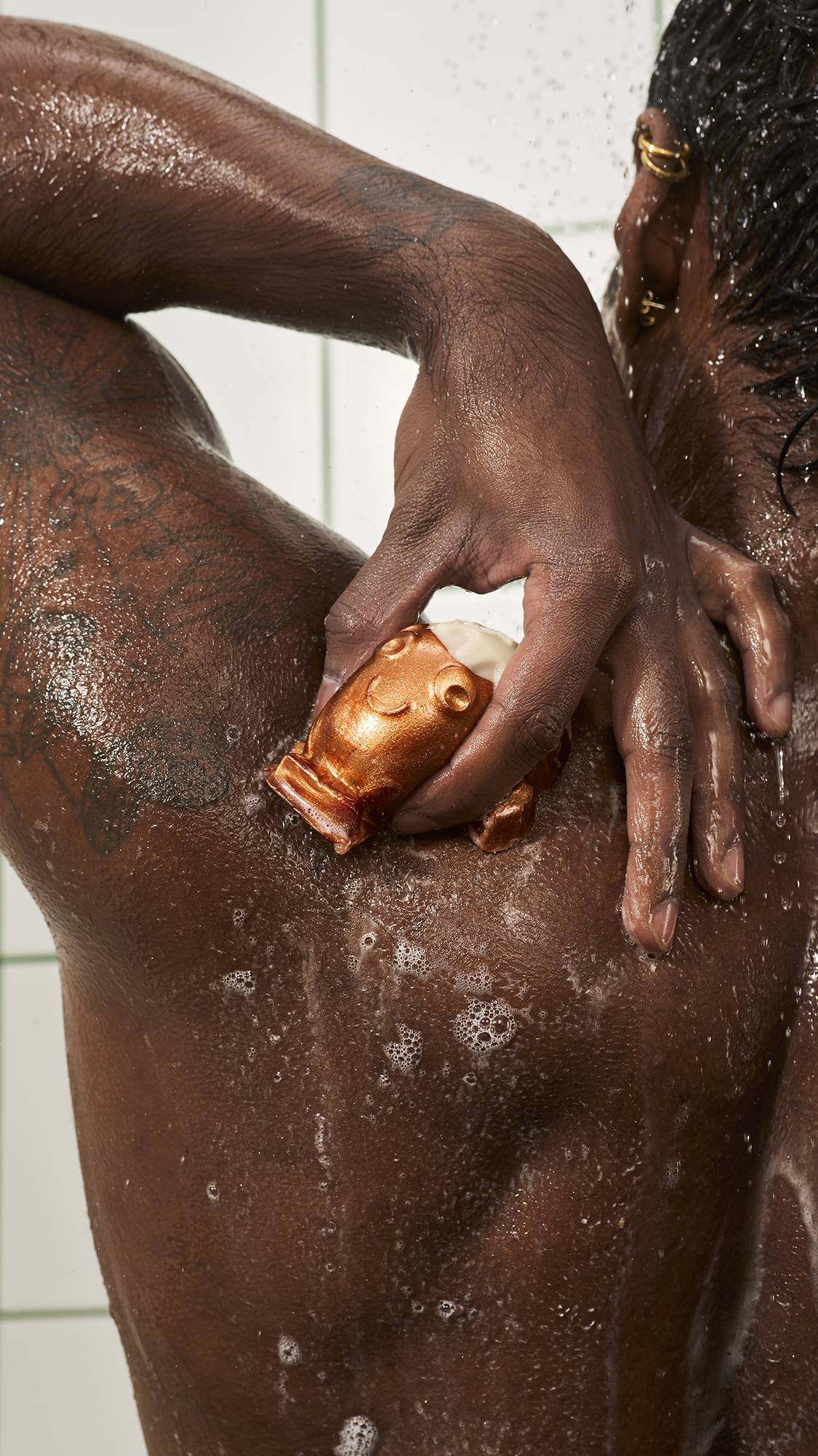 A person washes their wet shoulder with a bronze Lush "Christmas Pud" character jelly, spreading light soapy suds across their back in the shower against a white tiled wall.