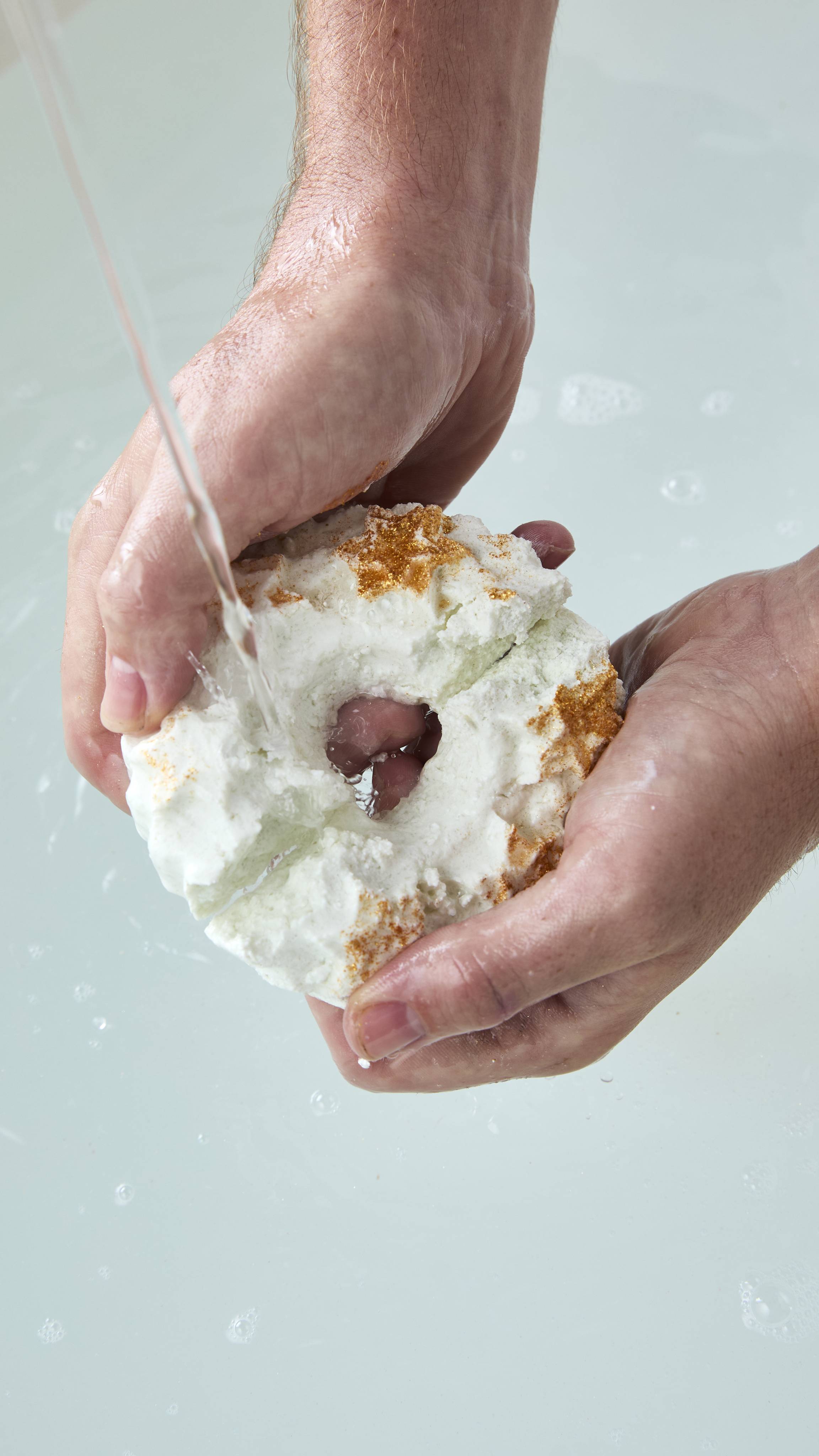 Two hands hold a white wreath-shaped bubble bar topped with gold stars under a running stream of tap water over clear bathwater, beginning to crumble and dissolve.
