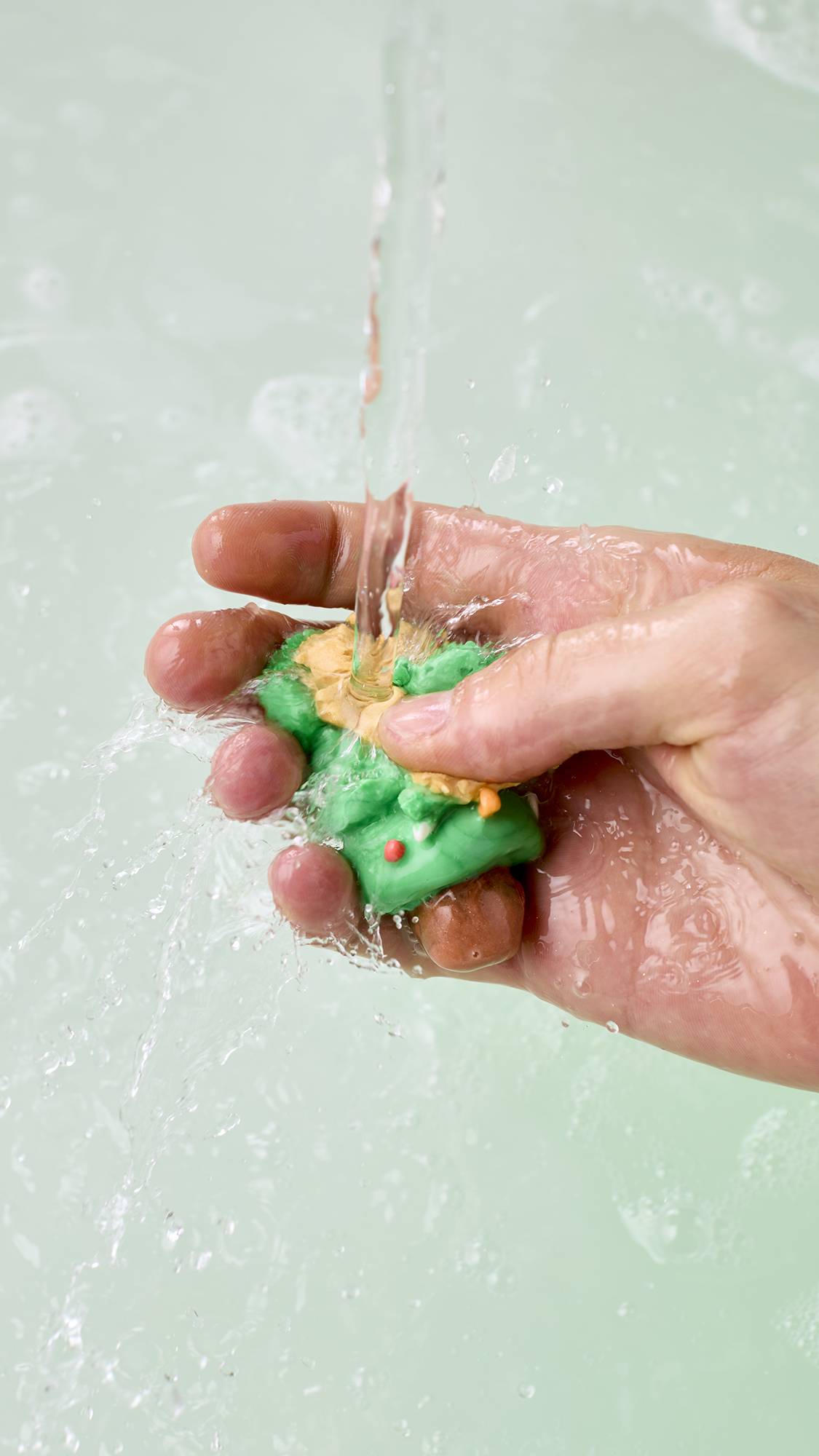 A hand squashes a green Christmas tree-moulded product with a yellow star under a stream of running tap water, crumbling it into clear bathwater as water splashes over the hand and product.