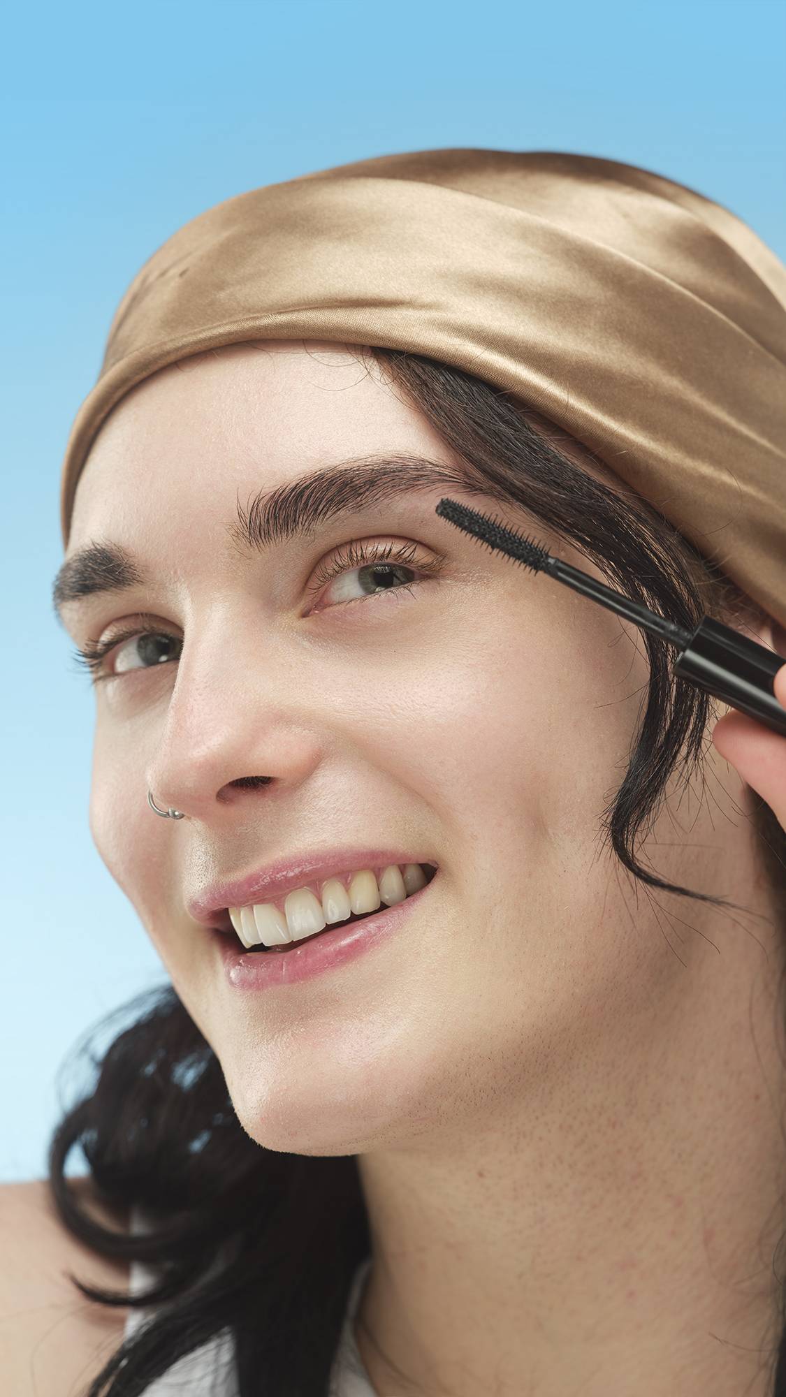 A person wearing a satin gold headband and a nose ring smiles while applying the Circus Lashes ~ Mascara to their eyebrows with the mascara wand. The product is being used to define the brows, against a bright blue background.