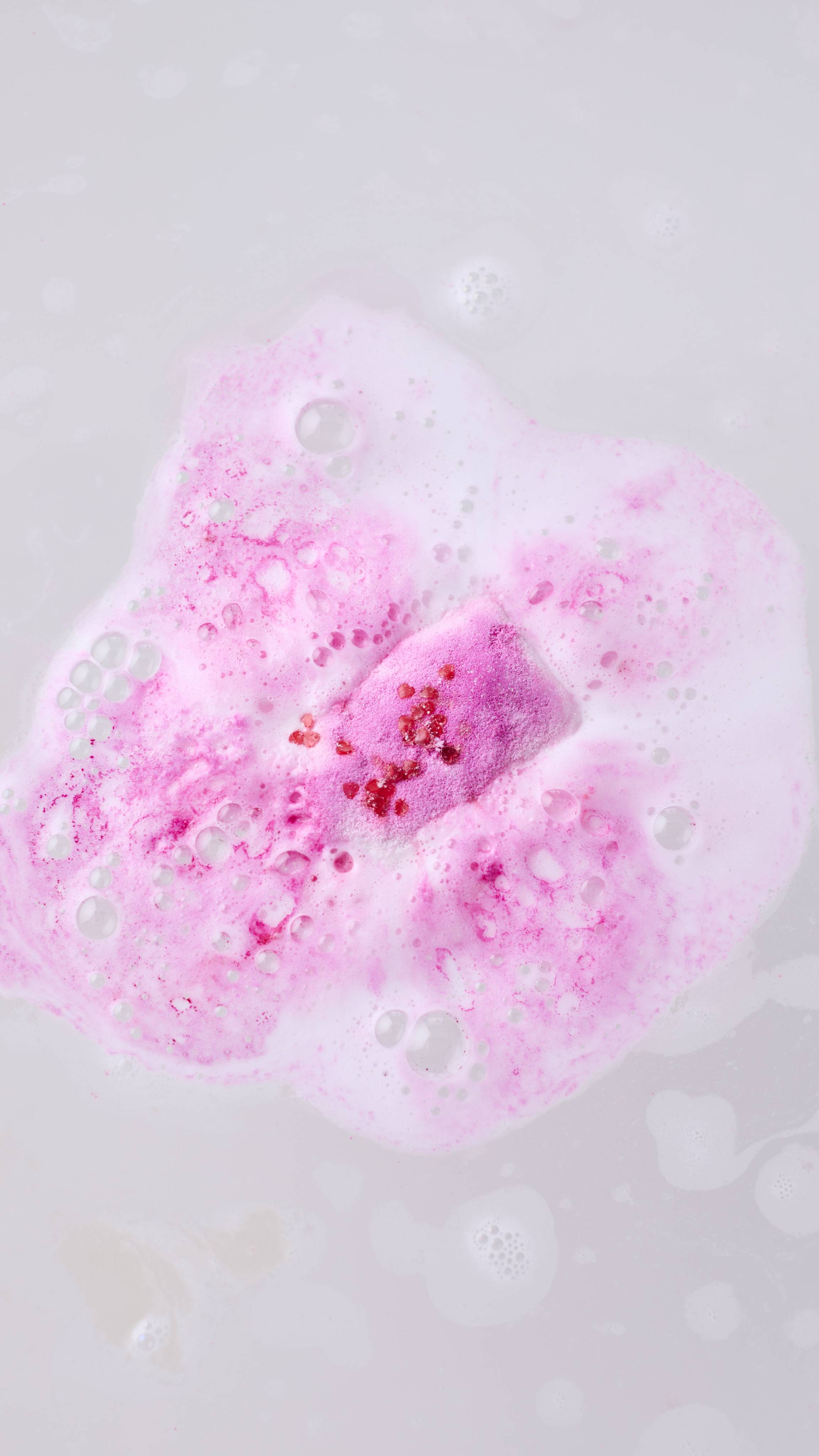 A top-down view shows a pink bath melt dissolving in milky water, releasing a cloud of vibrant pink foam topped with small red crystal sprinkles.