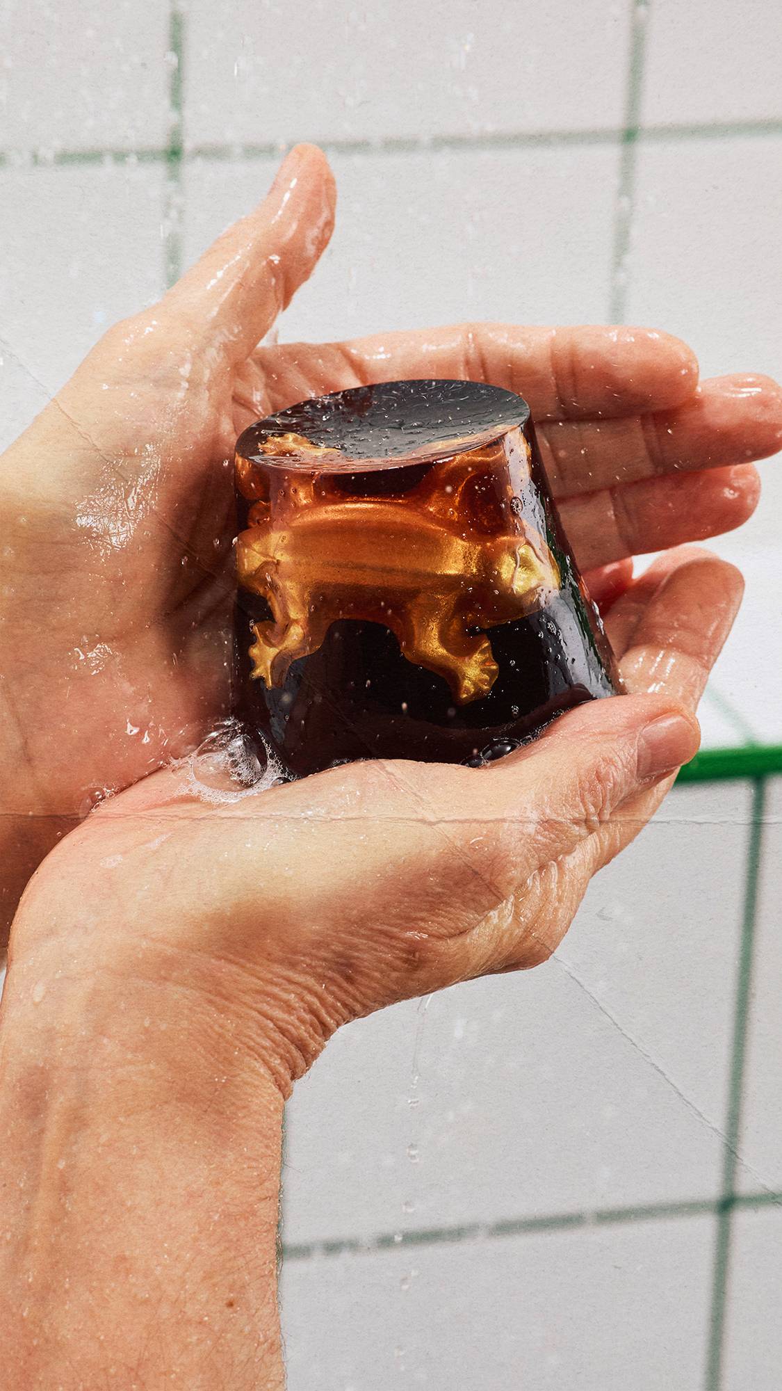 Two hands hold a wet, dark, translucent trapezoidal shower jelly under running shower water. A golden lizard gummy is embedded within the jelly. White square wall tiles with green grout lines form the background.