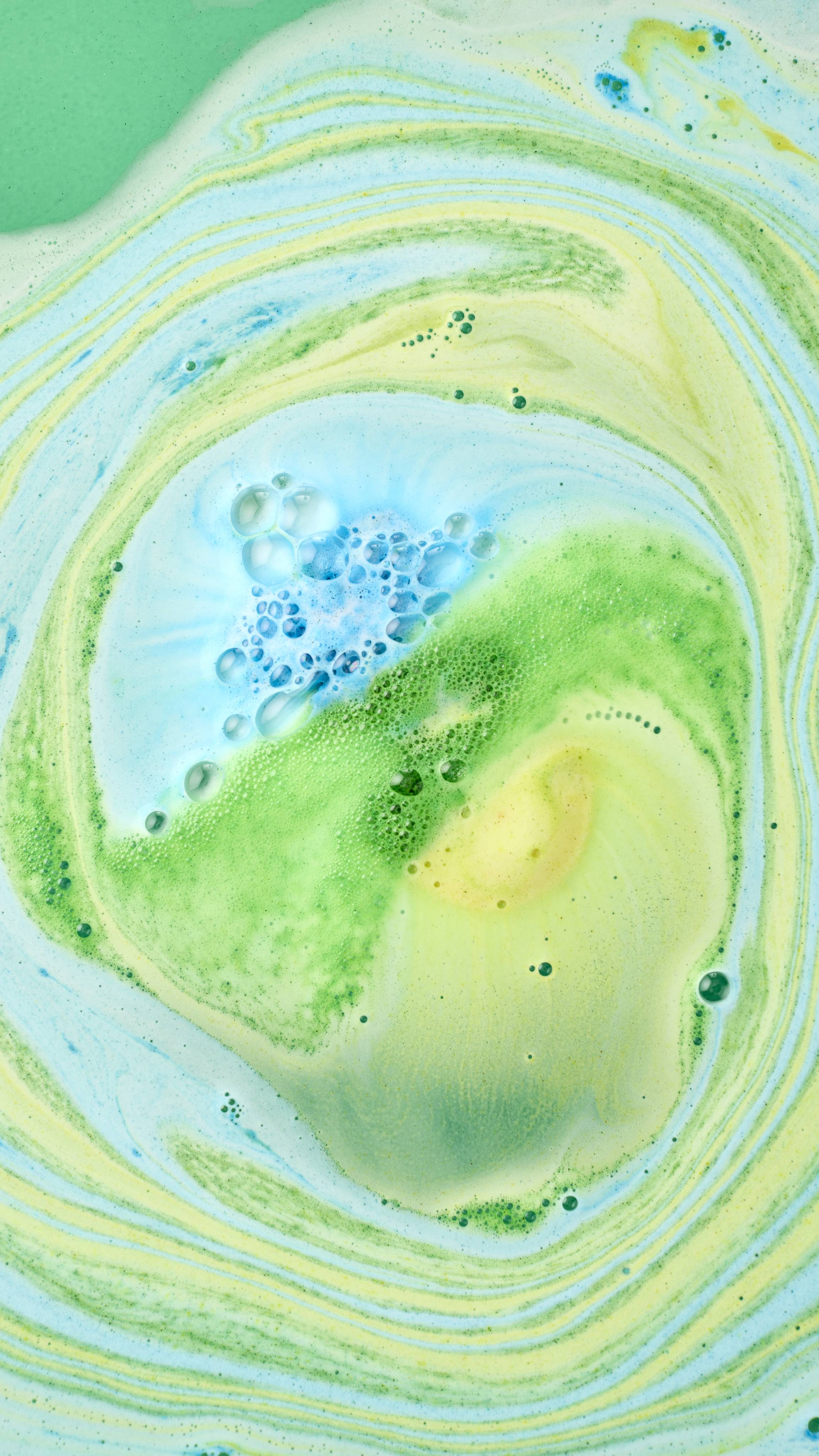 Vibrant blue, green and yellow foam swirls together in a bathtub from the "DAD" bath bomb, creating a marbled pattern of green, blue and yellow as the product continues to dissolve and blend into the water.
