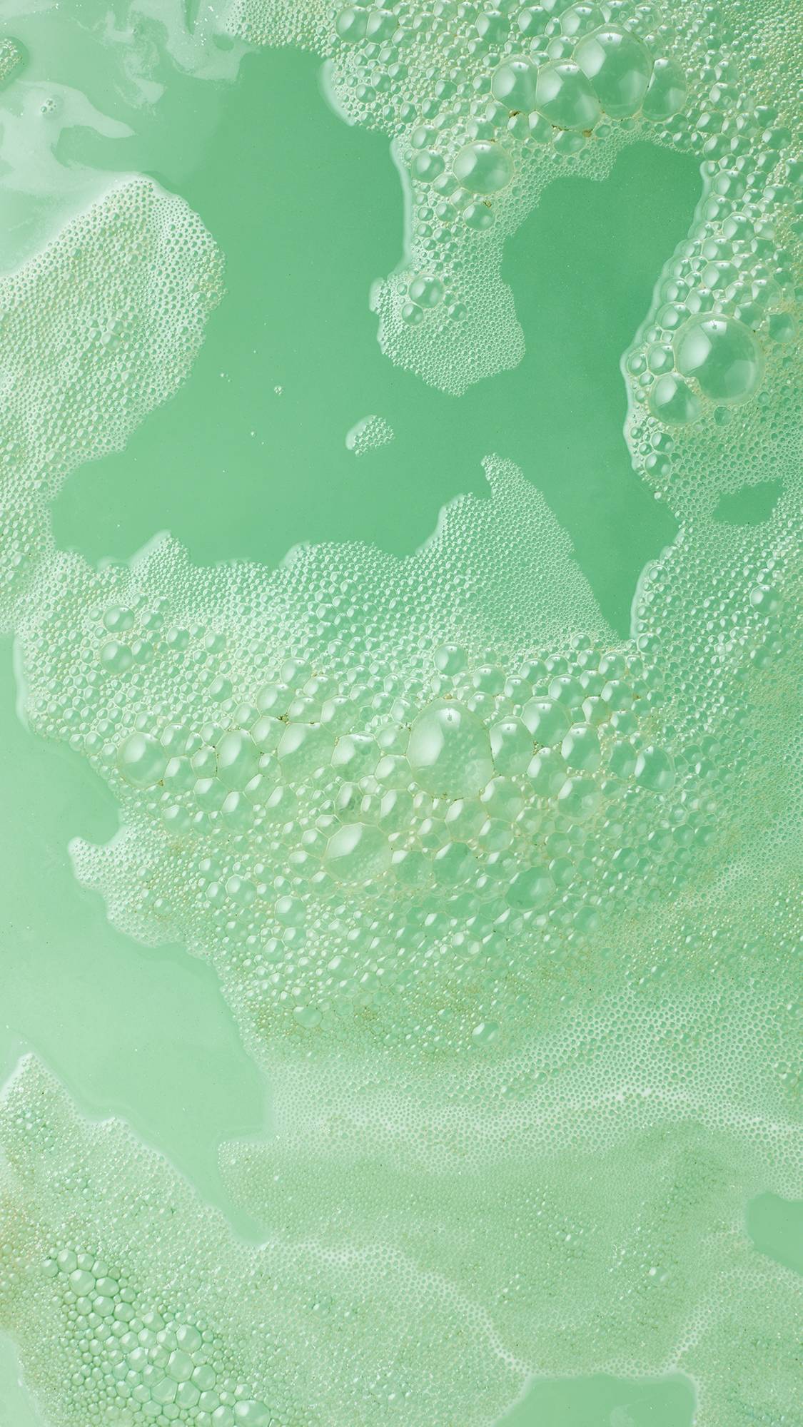 Thick, seafoam-green foam drifts across pale mint bathwater, forming soft textured bubbles and shimmering, pale-green swirls as a foot bomb fully dissolves.