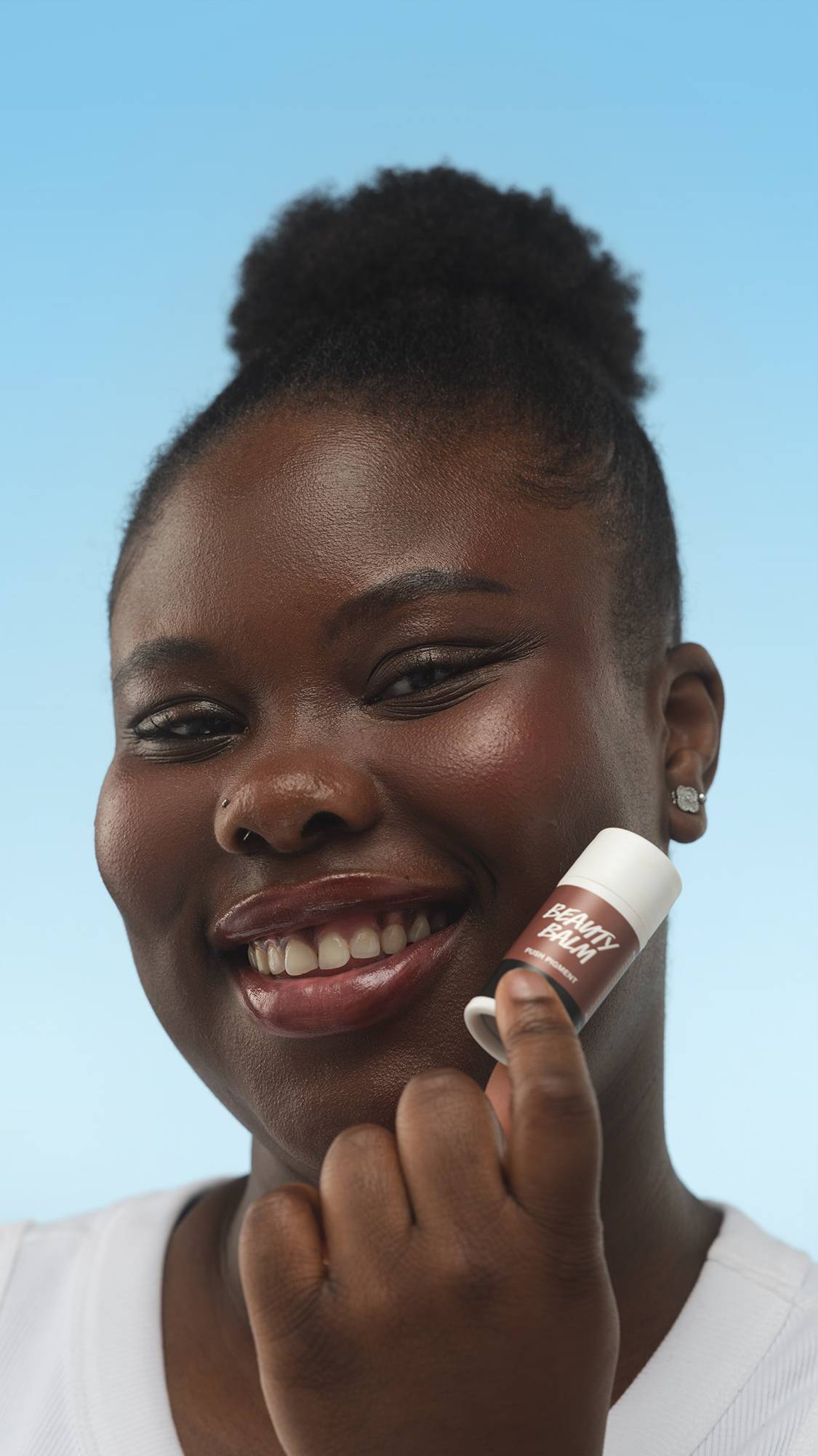 A close-up vertical portrait of a smiling person with deep skin, holding the white tube of Dark Cool Beauty Balm near their cheek. Their dark hair is in a bun, set against a bright blue background.