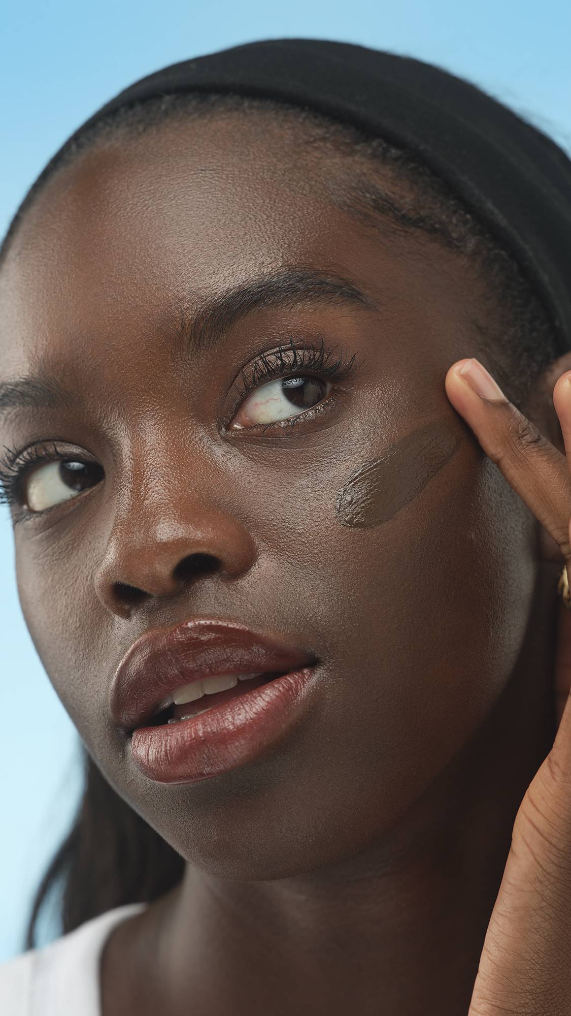 A close-up vertical portrait of a person with deep skin, wearing a black headband, looking up and right, while applying a creamy dark brown swatch of Dark Neutral Skinwear to their cheek with a finger. The background is a bright blue.