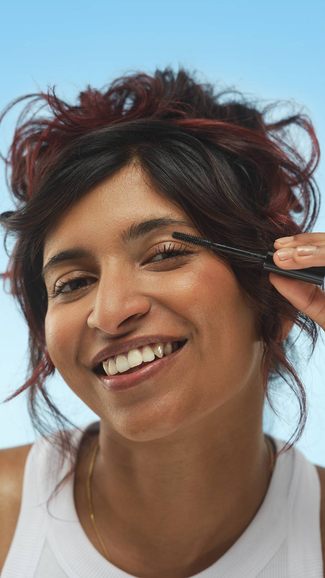 A person with dark hair and red highlights, wearing a white ribbed top, smiles while applying Eyes Right ~ Mascara to their upper eyelashes with the black wand. They have a subtle shimmer on their cheek, set against a bright blue background.