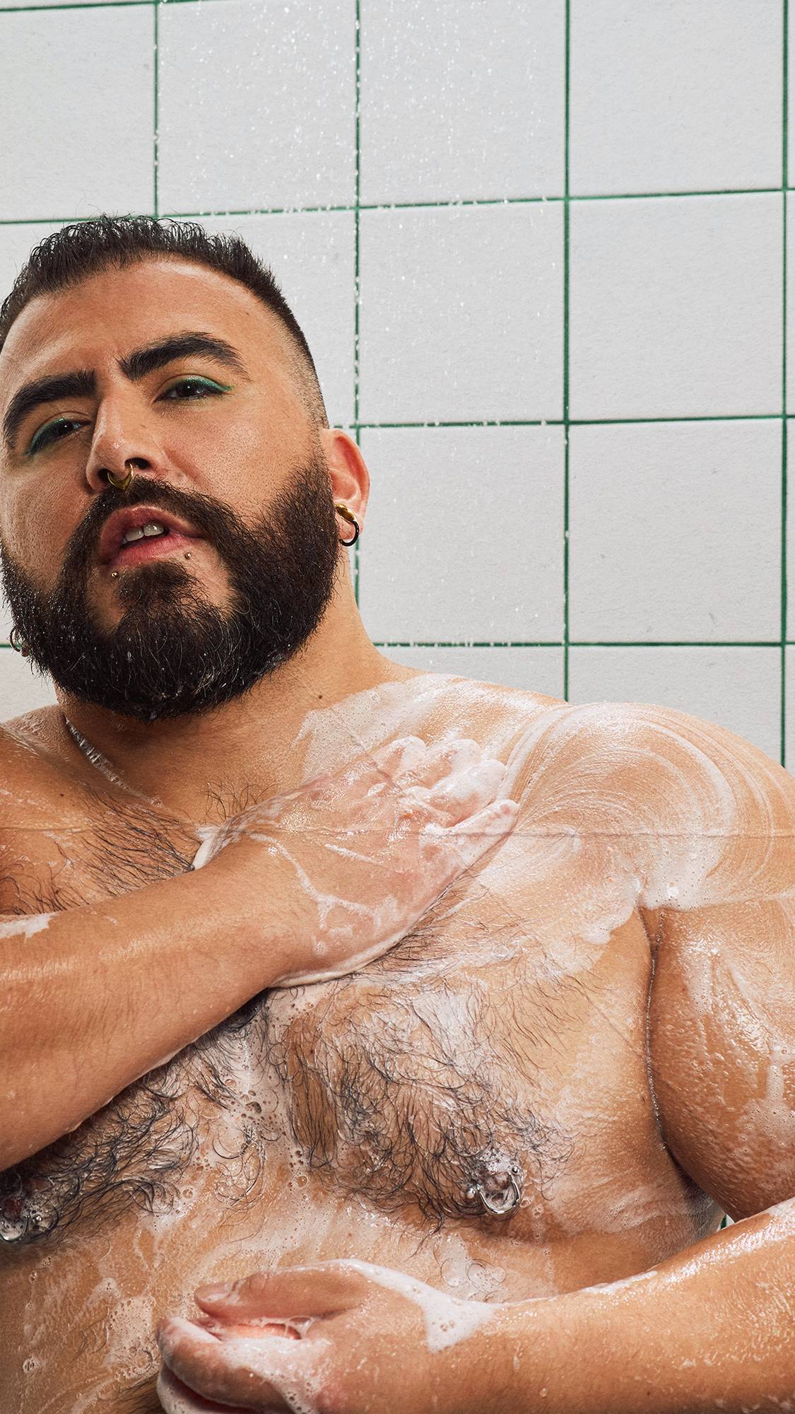 A bearded model with green eyeliner lathers creamy white soap across their chest and shoulders in the shower, standing against a backdrop of white tiles with green grout.