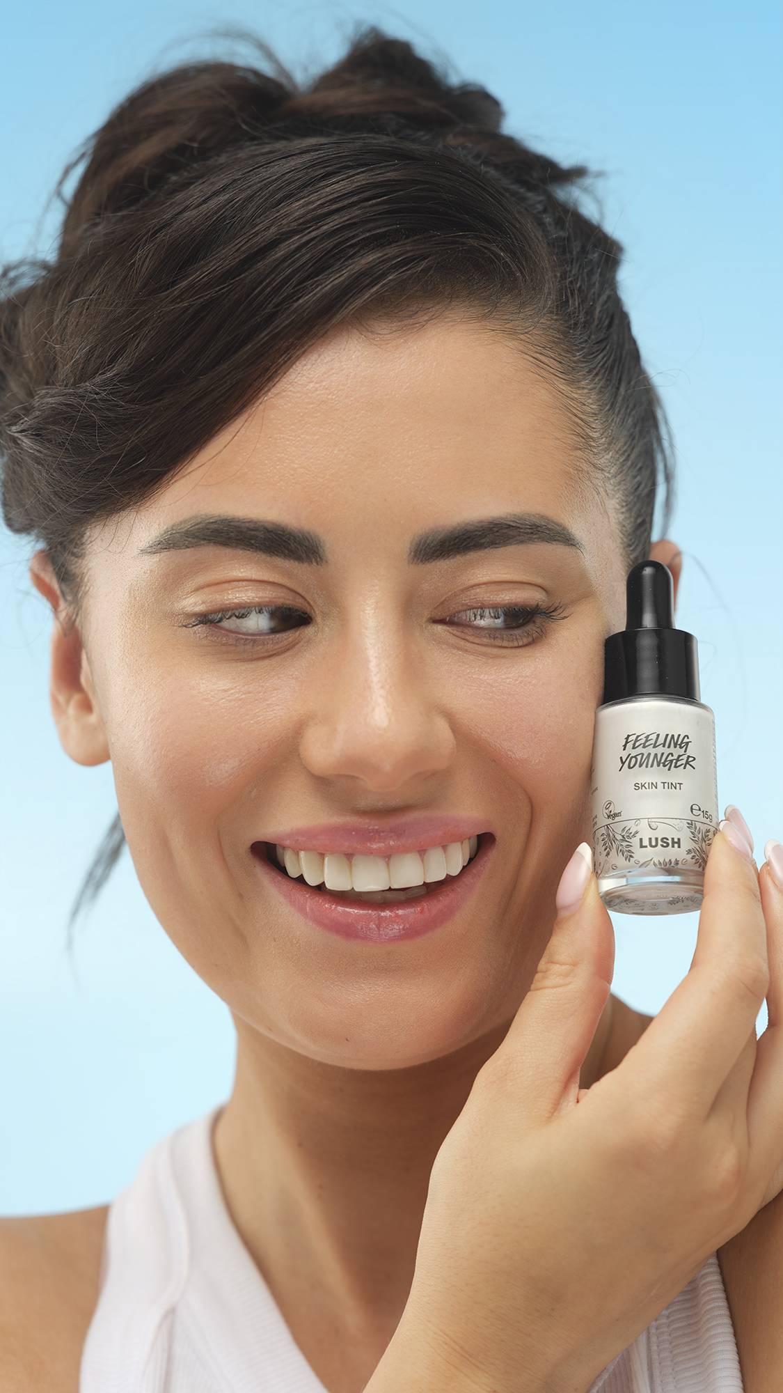 A close-up vertical portrait of a smiling person with medium skin, dark hair pulled up, holding the dropper bottle of LUSH Feeling Younger Skin Tint next to their face. Their skin has a luminous finish, set against a bright blue background.