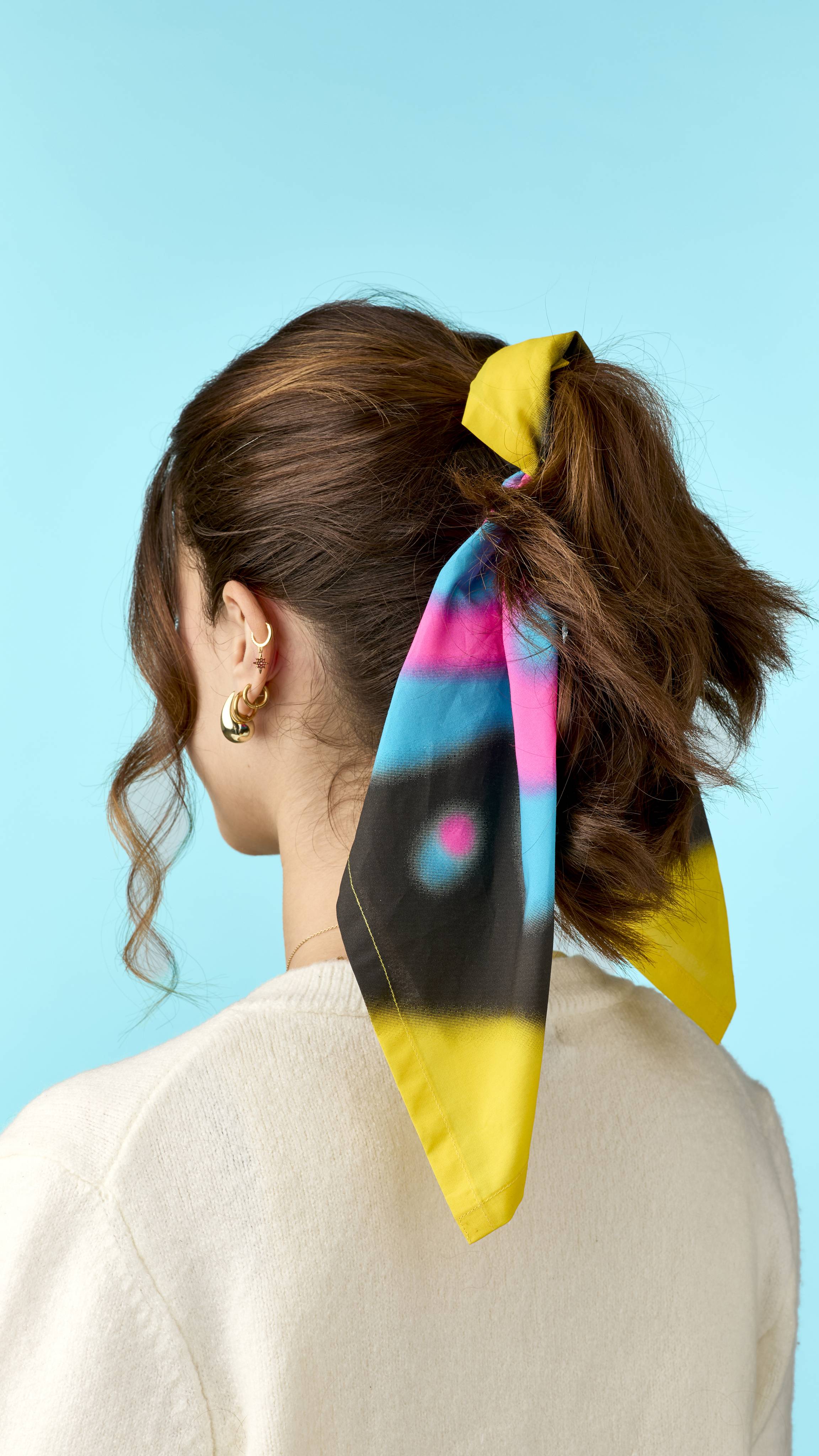 A person with brown hair tied in a high ponytail wearing a colourful patterned fabric knot wrap scarf in yellow, pink, blue, and black, styled with gold earrings against a light blue background.