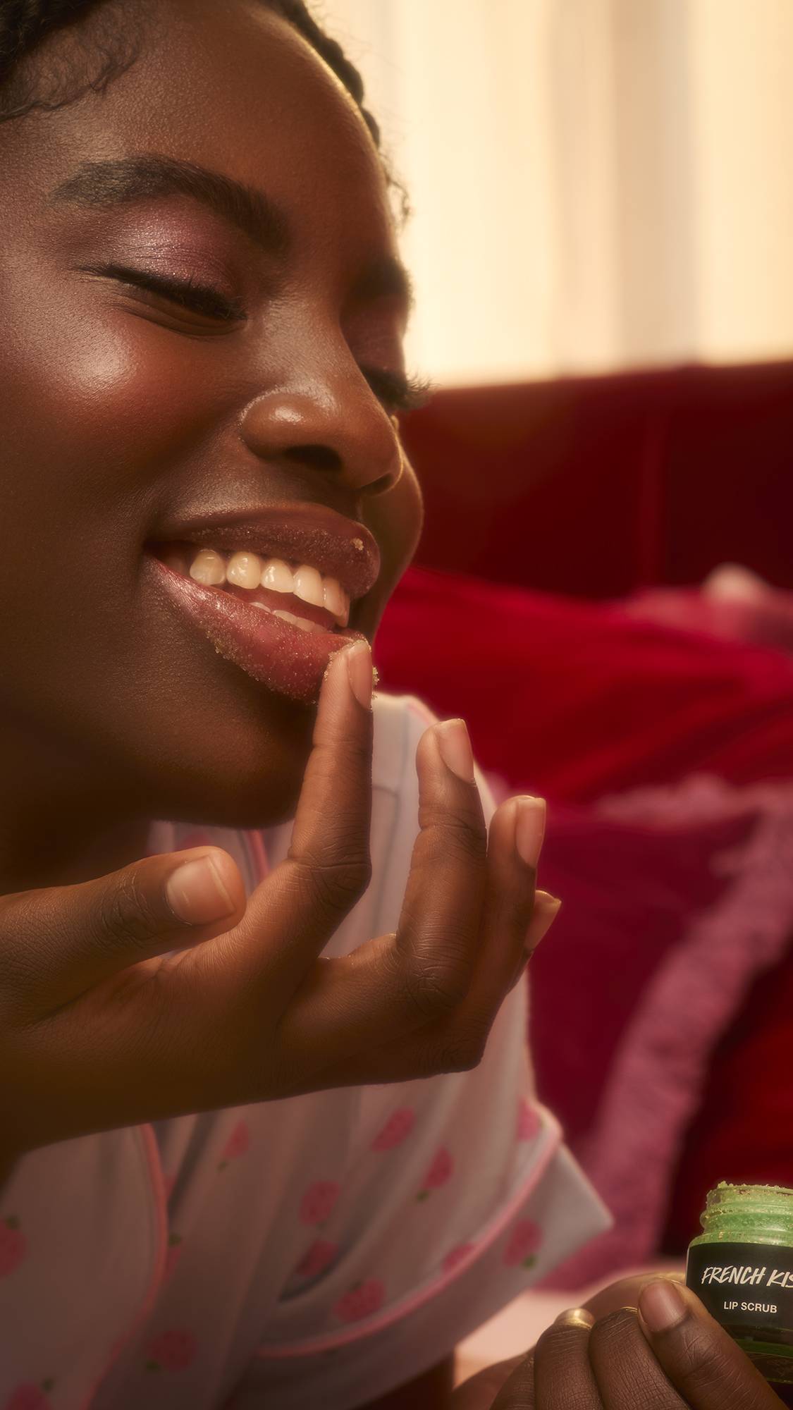 A close-up image shows the grainy, sparkling texture of Lush "French Kiss" lip scrub. A person with dark skin uses a finger to apply the bright green, sugary paste to their lips.