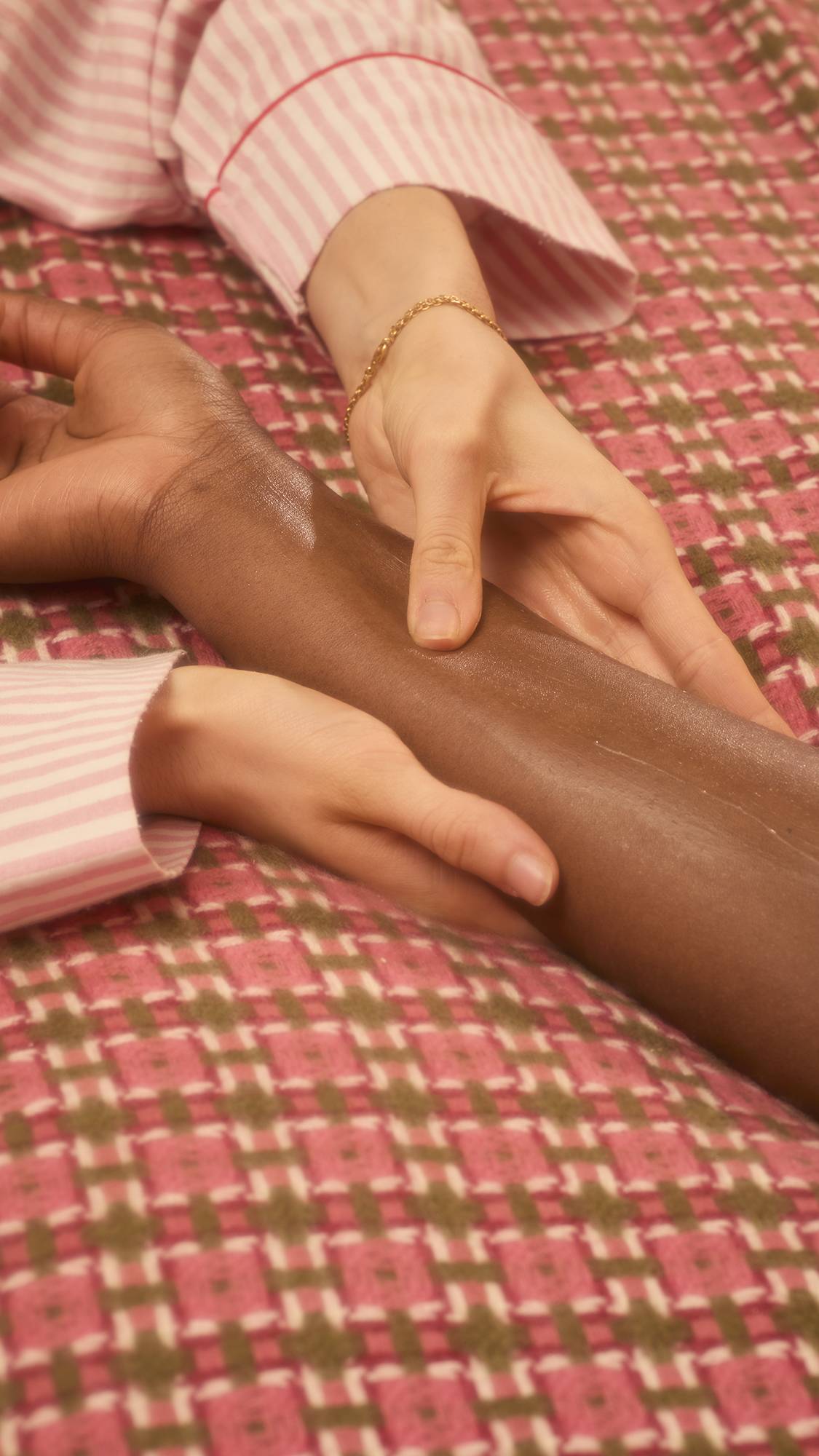 A person wearing striped pyjamas uses both hands to massage a cream-coloured lotion into someone else's forearm using the Gorgeous massage bar. The pair are positioned over a red, brown, and pink patterned bedspread.