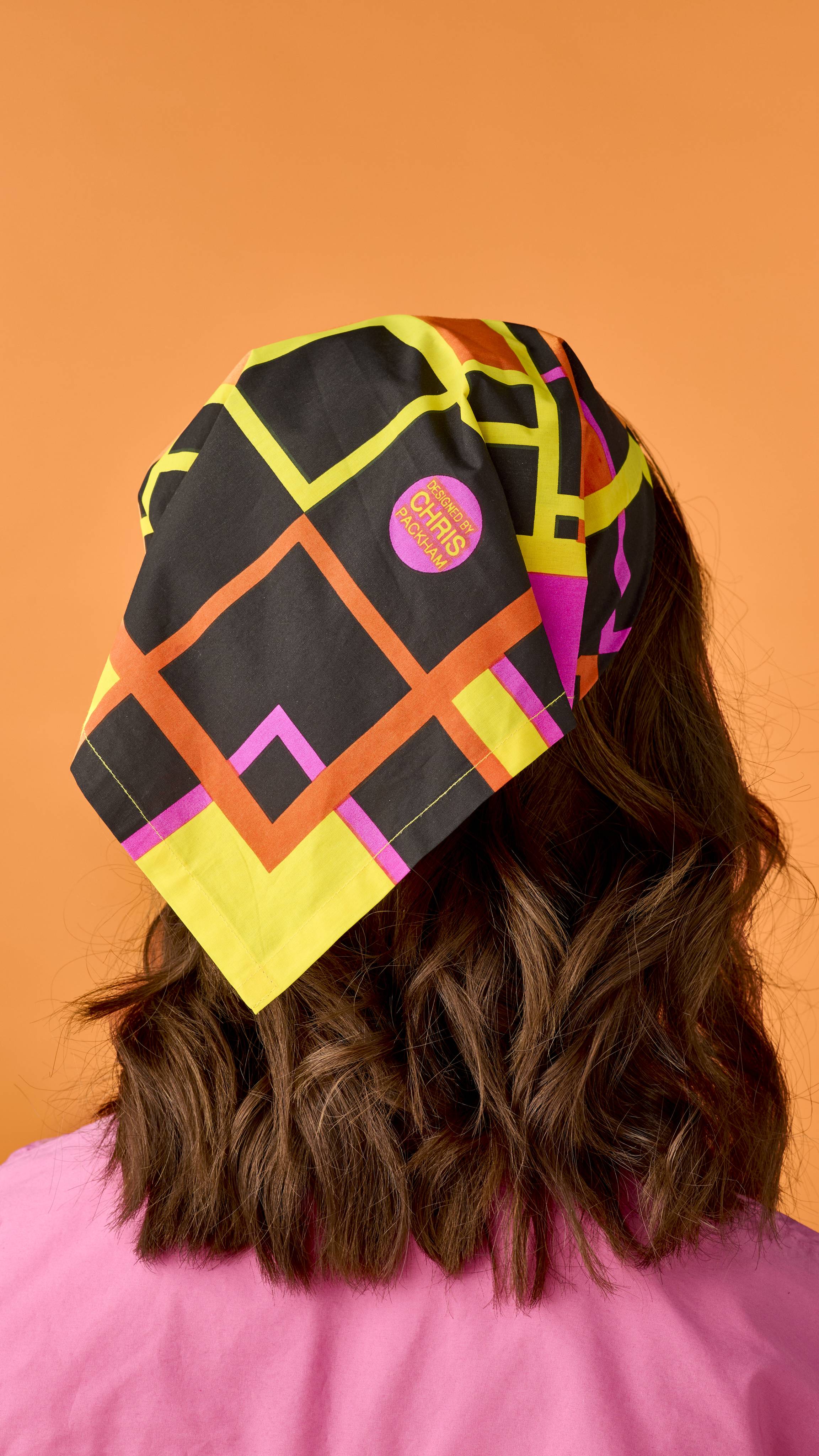 A person with brown wavy hair viewed from behind, wearing a headscarf featuring a black, yellow, orange, and pink geometric pattern, detailed with a circular pink stamp that reads "DESIGNED BY CHRIS PACKHAM".