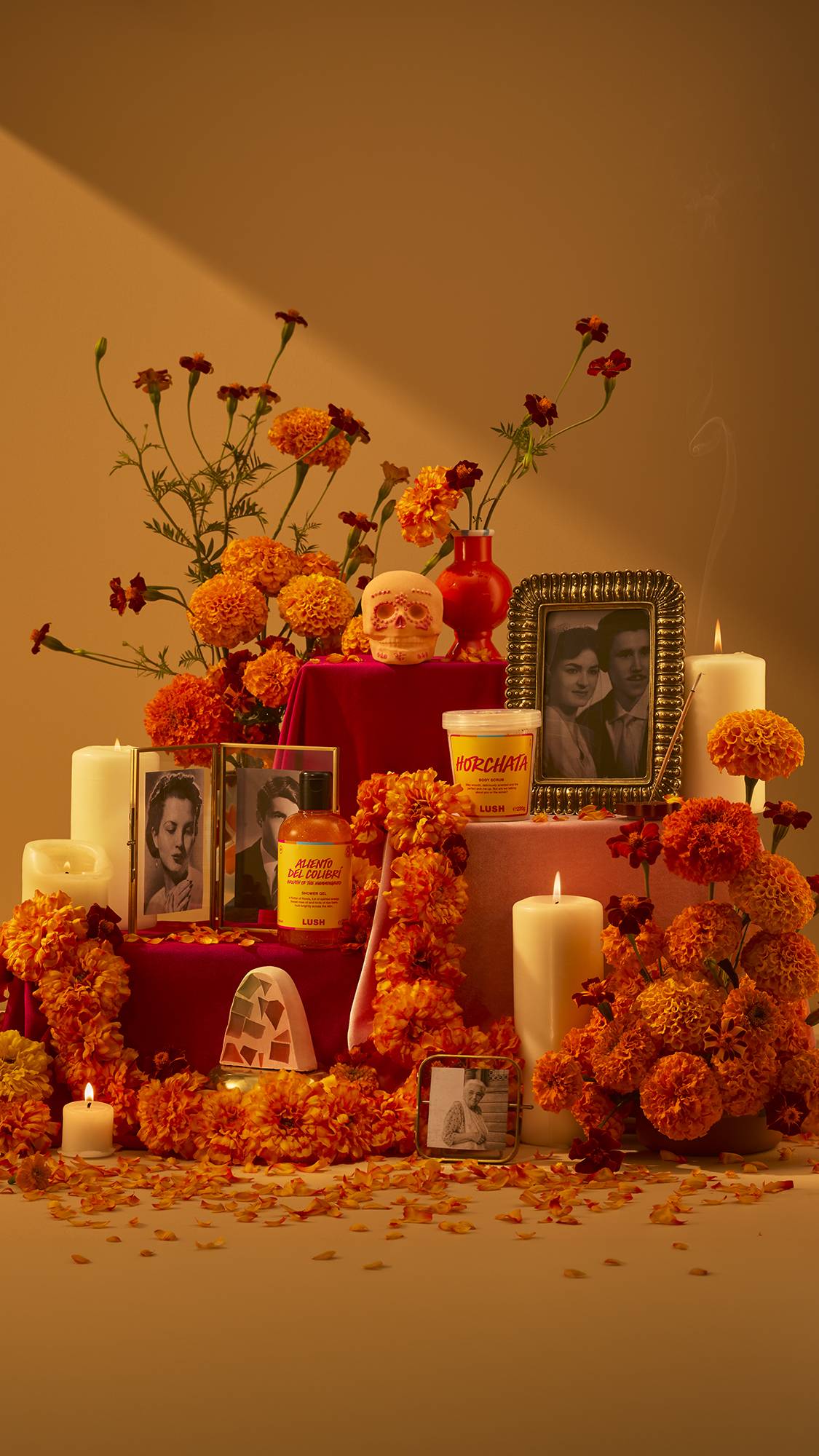 A Día de Muertos altar arrangement featuring orange marigolds, lit candles, framed photos, with the range of Lush products, including a skull bath bomb, shower gel, body scrub, soap, and a colourful knot-wrap gift.