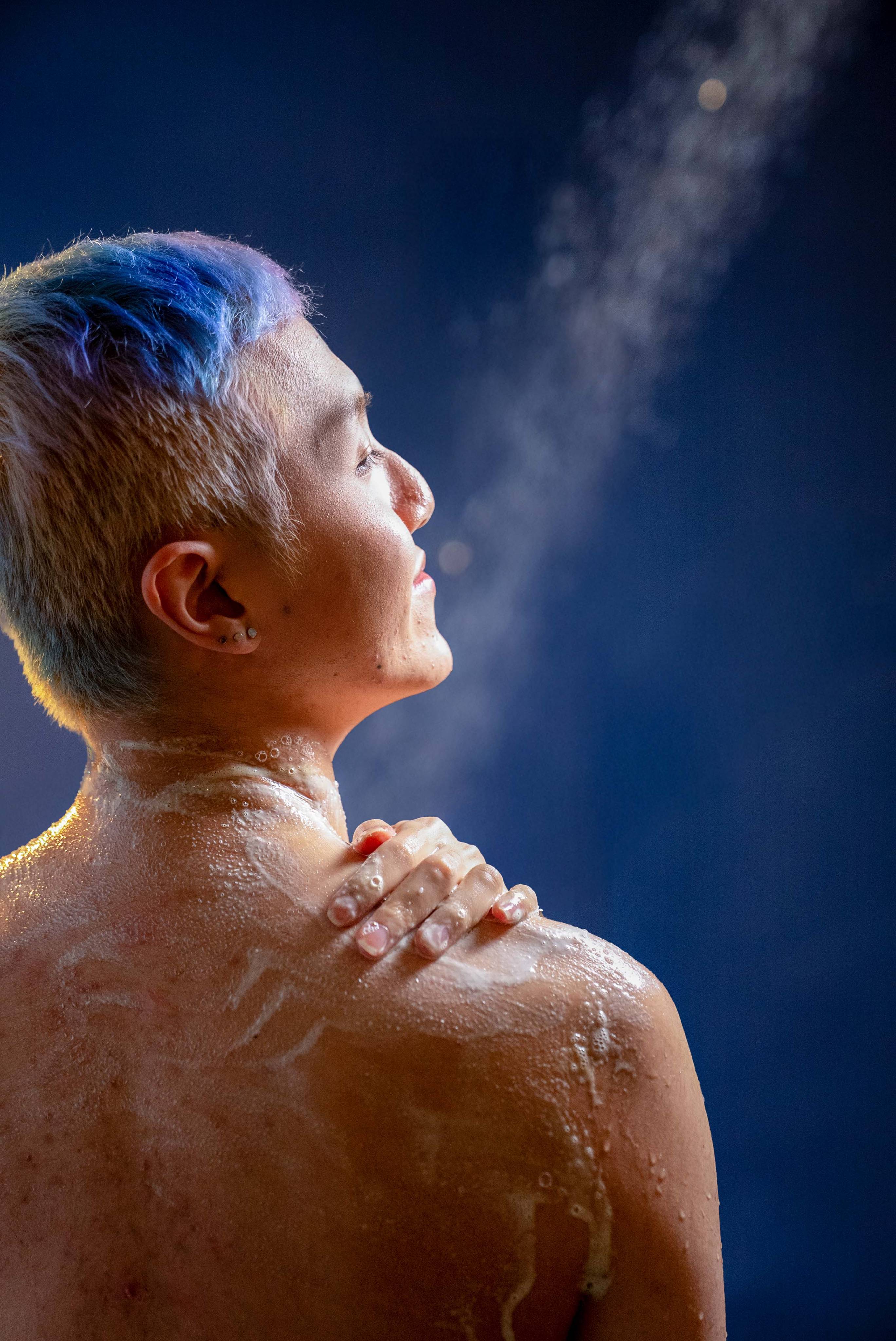 A person with short, blue and purple dyed hair looks up towards a stream of water in the shower. Their back and shoulder are covered in soapy white lather as they rest a hand on their shoulder against a dark blue background.