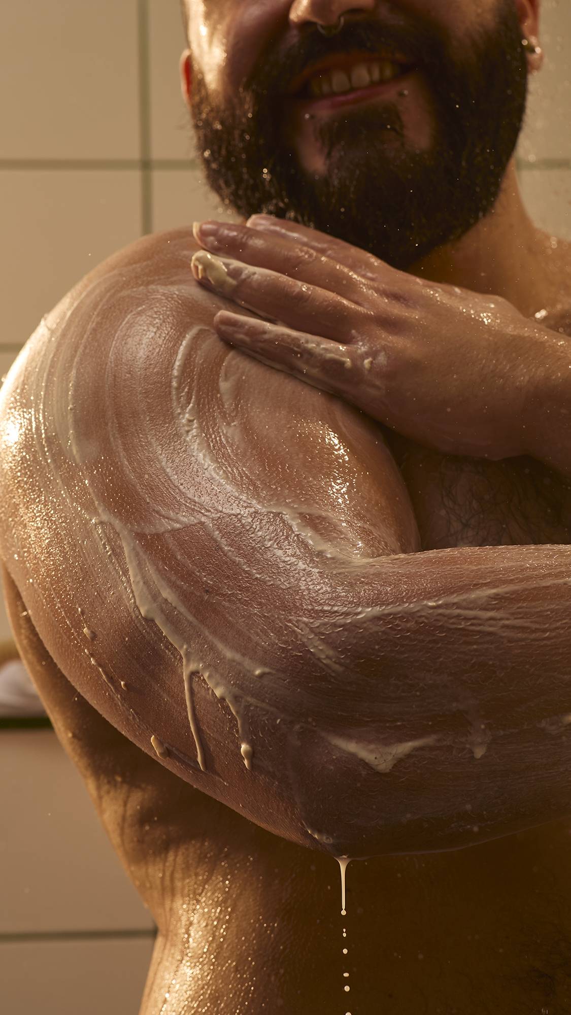 A bearded model with facial piercings smiles as they massage the Lush Día De Muertos "Horchata" creamy body scrub into their shoulder and chest under a warm spray of water in the shower.
