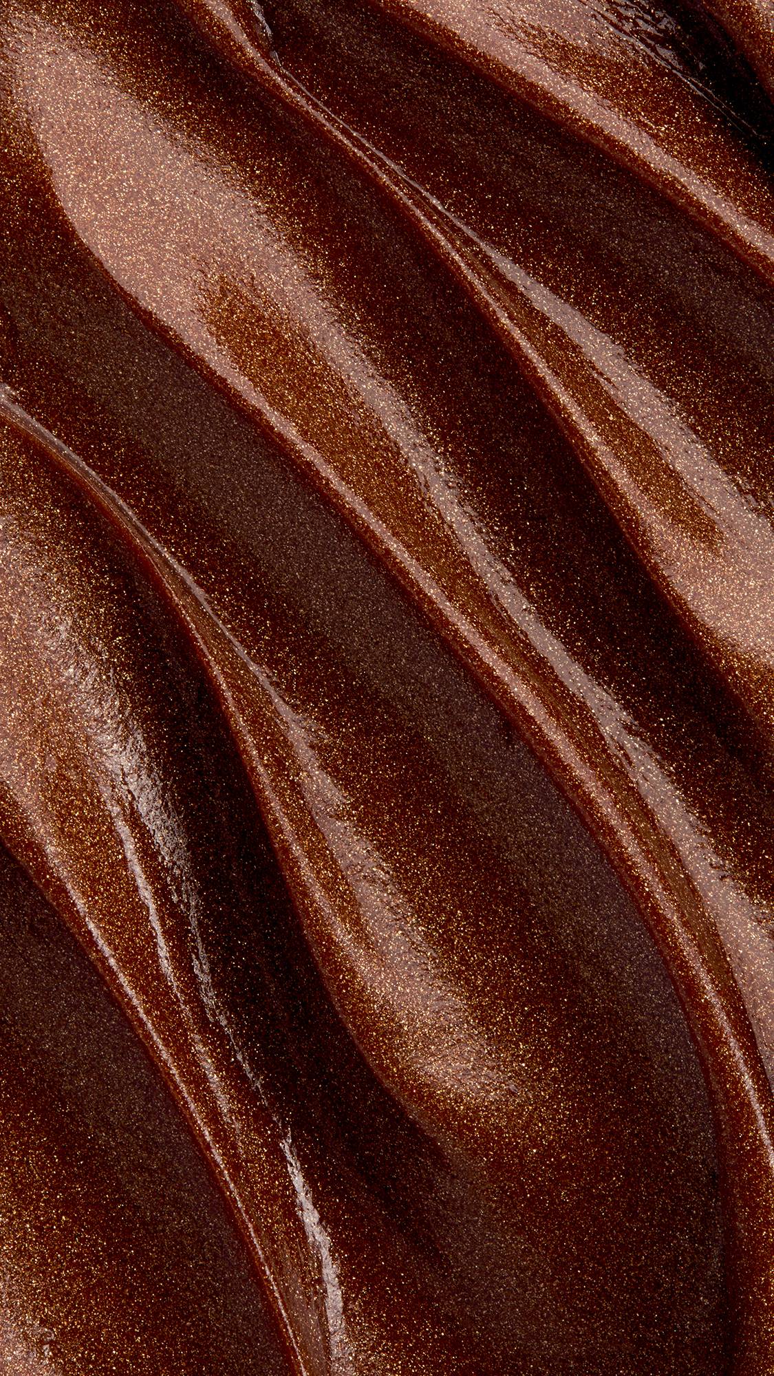 A macro close-up texture shot of rich, glossy bronze-brown lip mask spread in smooth diagonal swirls, filled with fine, shimmering copper glitter particles across the frame.