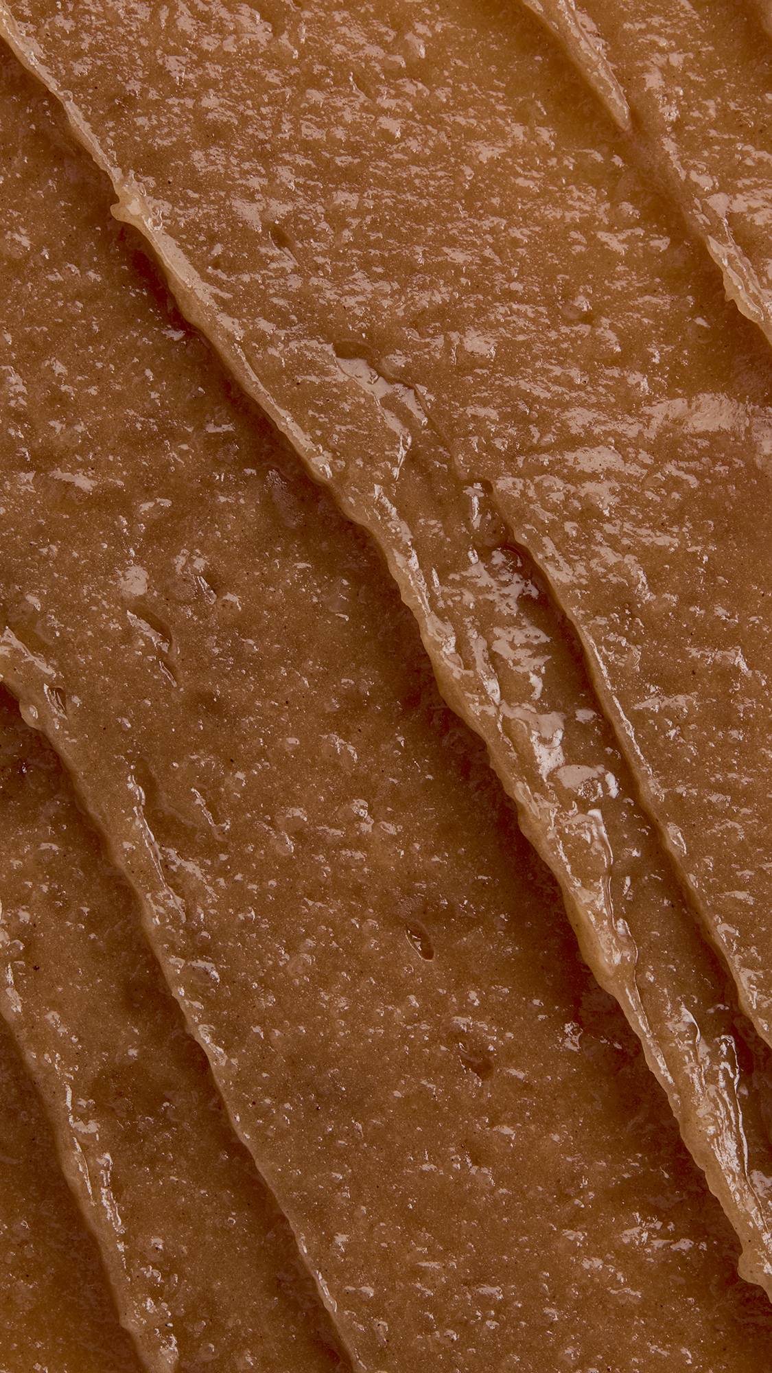 A macro close-up texture shot of rich, light-brown lip butter spread in thick, glossy diagonal ridges with a smooth, moisturizing finish across the frame.
