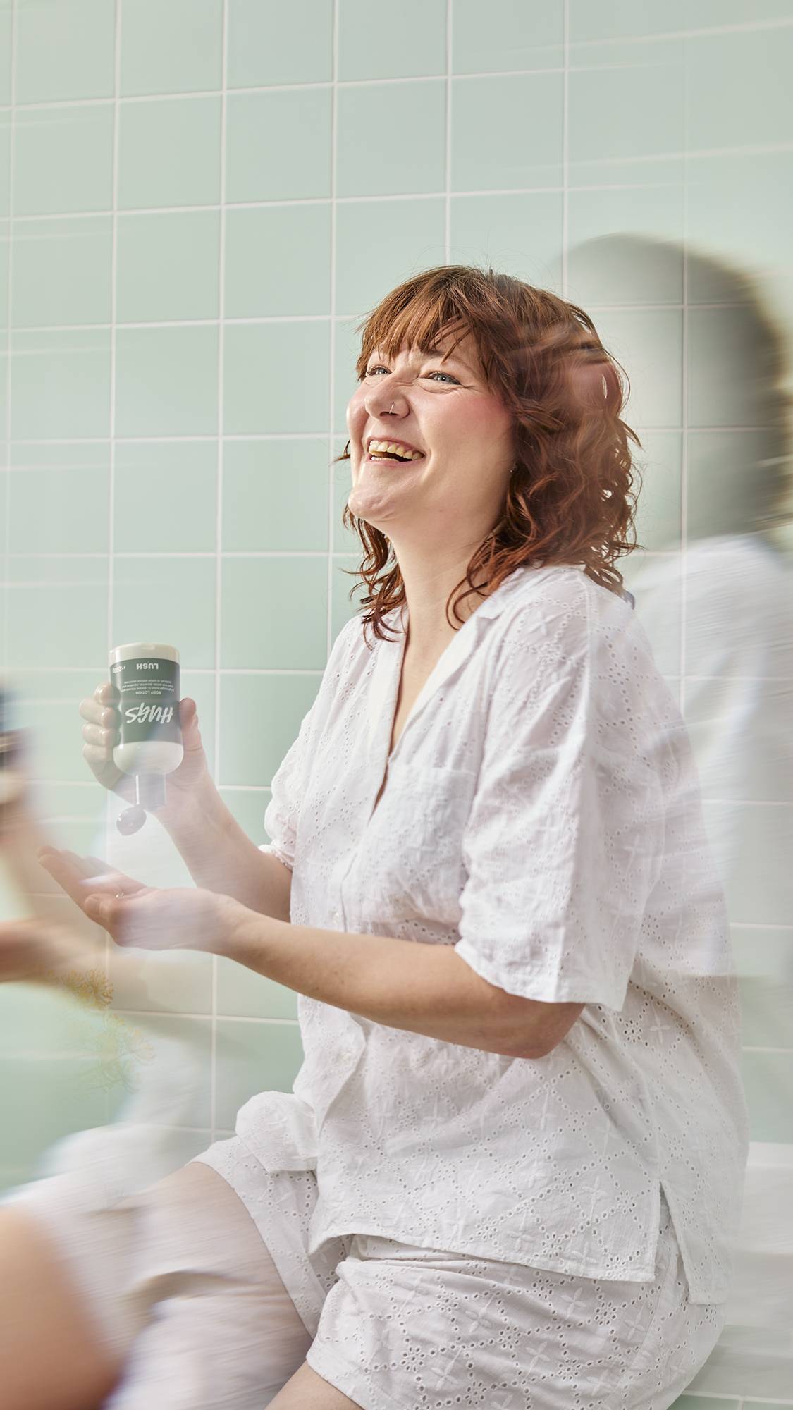 A model with wavy auburn hair laughs joyfully while sitting in a bathroom with pale green tiles. They wear white pyjamas and hold a bottle of Lush "Hugs" Body Lotion, ready to dispense it into their open palm.