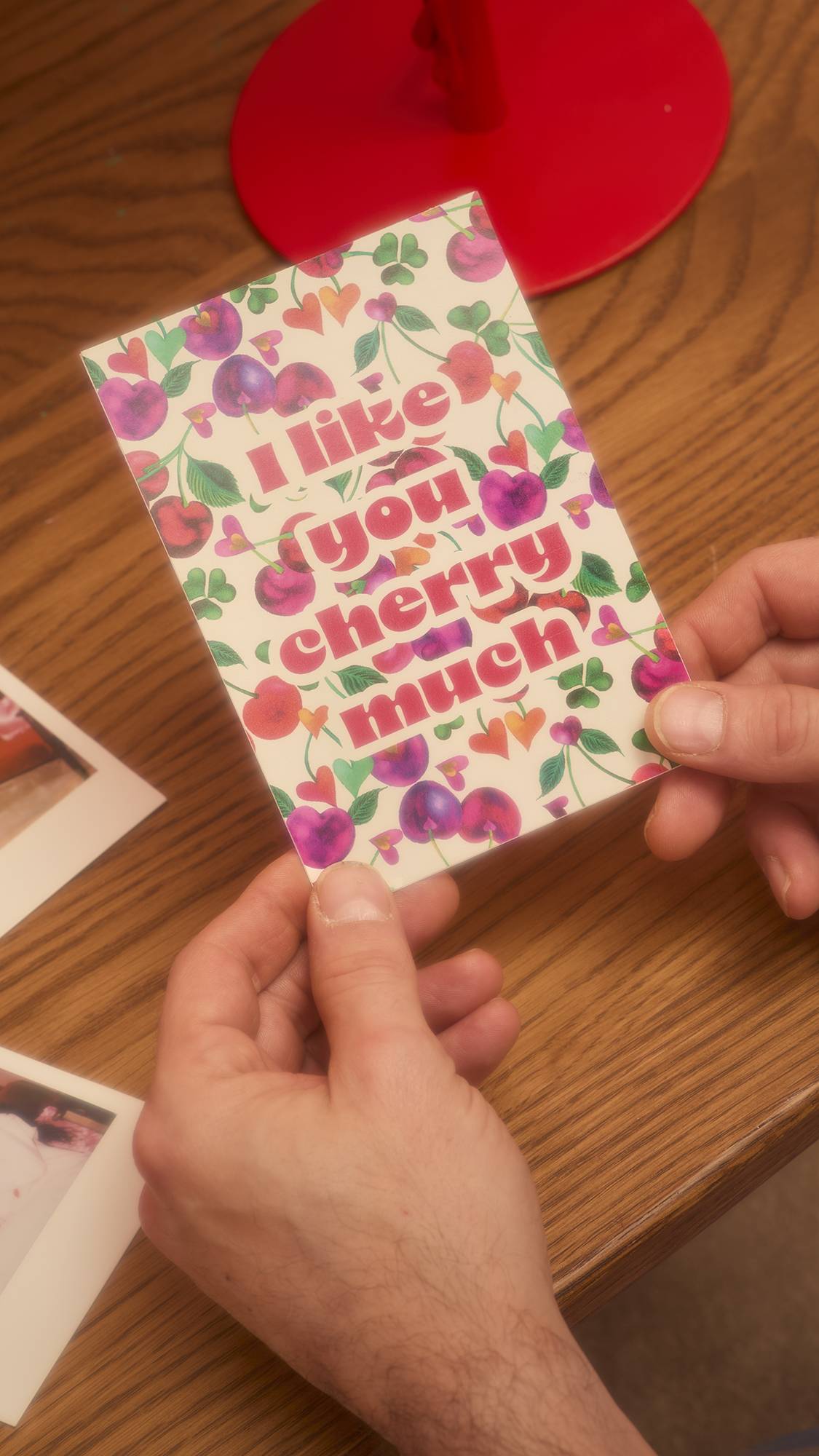 Hands hold the "I Like You Cherry Much" card, showcasing the heart-shaped cherry pattern and glittery red text. The card rests above a wooden table scattered with Polaroid photos.