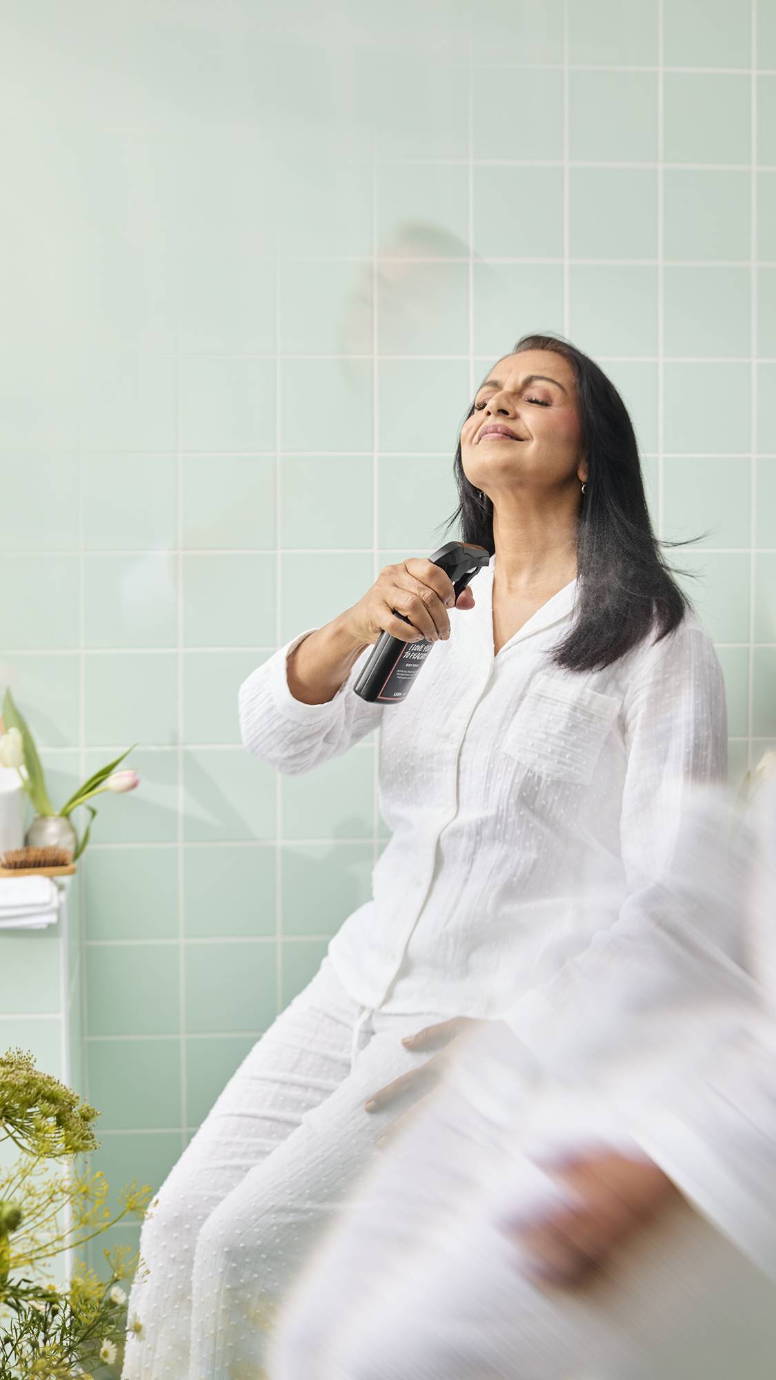 A model with long black hair sits in a bathroom with pale green tiles, wearing white pyjamas. They hold a bottle of Lush "I Love You To Peaches" Body Spray and mistily sprays it towards their neck with a peaceful, eyes-closed expression.