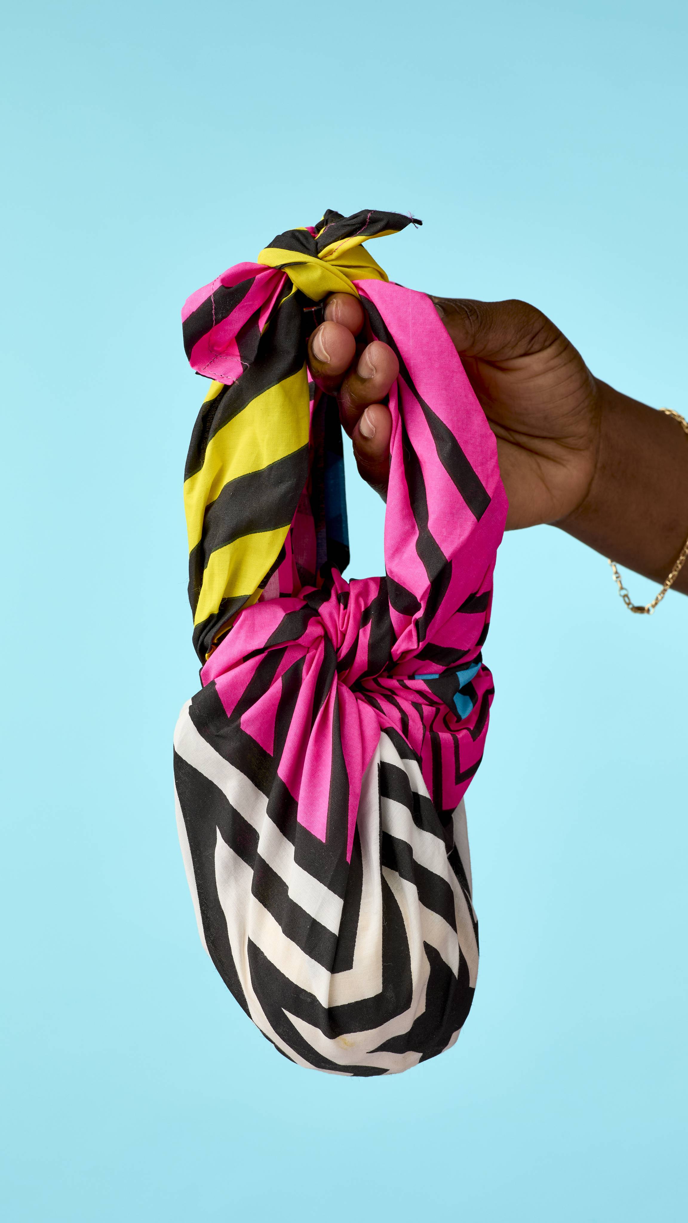 A hand holds up a knot-wrapped fabric bag by its tied top handle against a solid bright blue background, featuring bold geometric stripes in pink, black, white, and yellow.