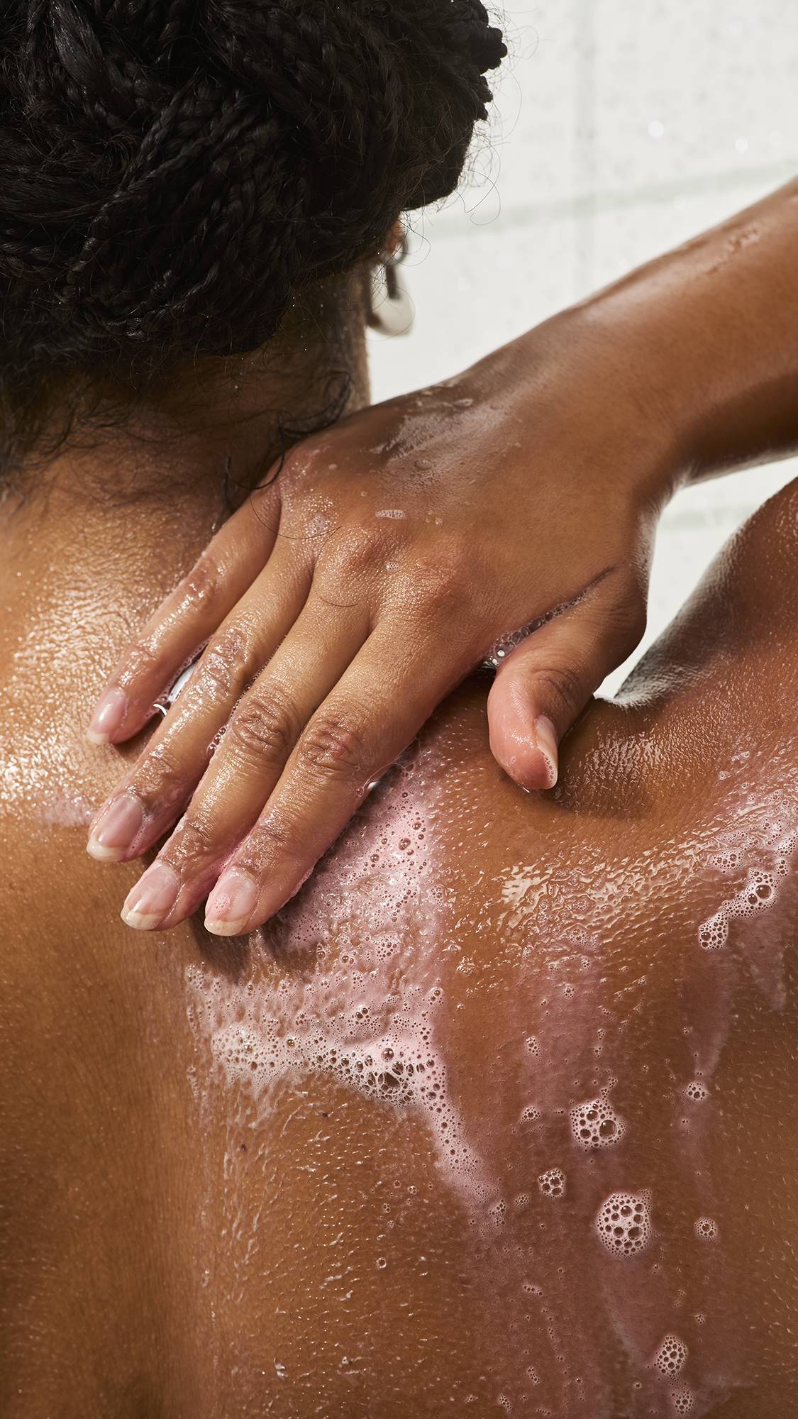 A close-up of a person with braided hair washing their upper back and shoulder, smoothing soapy white lather across wet skin under running shower water.