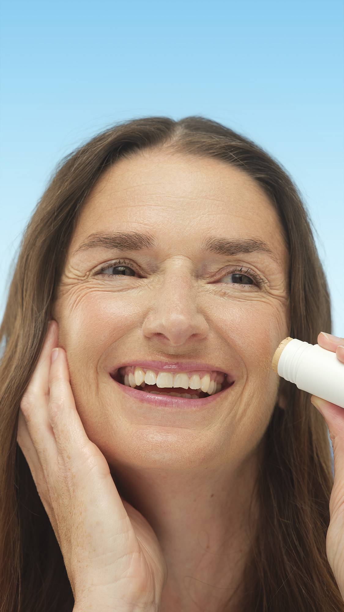 A close-up vertical portrait of a smiling person with warm-toned skin, applying the stick of Light Medium Warm Beauty Balm to their cheek. They have long brown hair, set against a bright blue background.