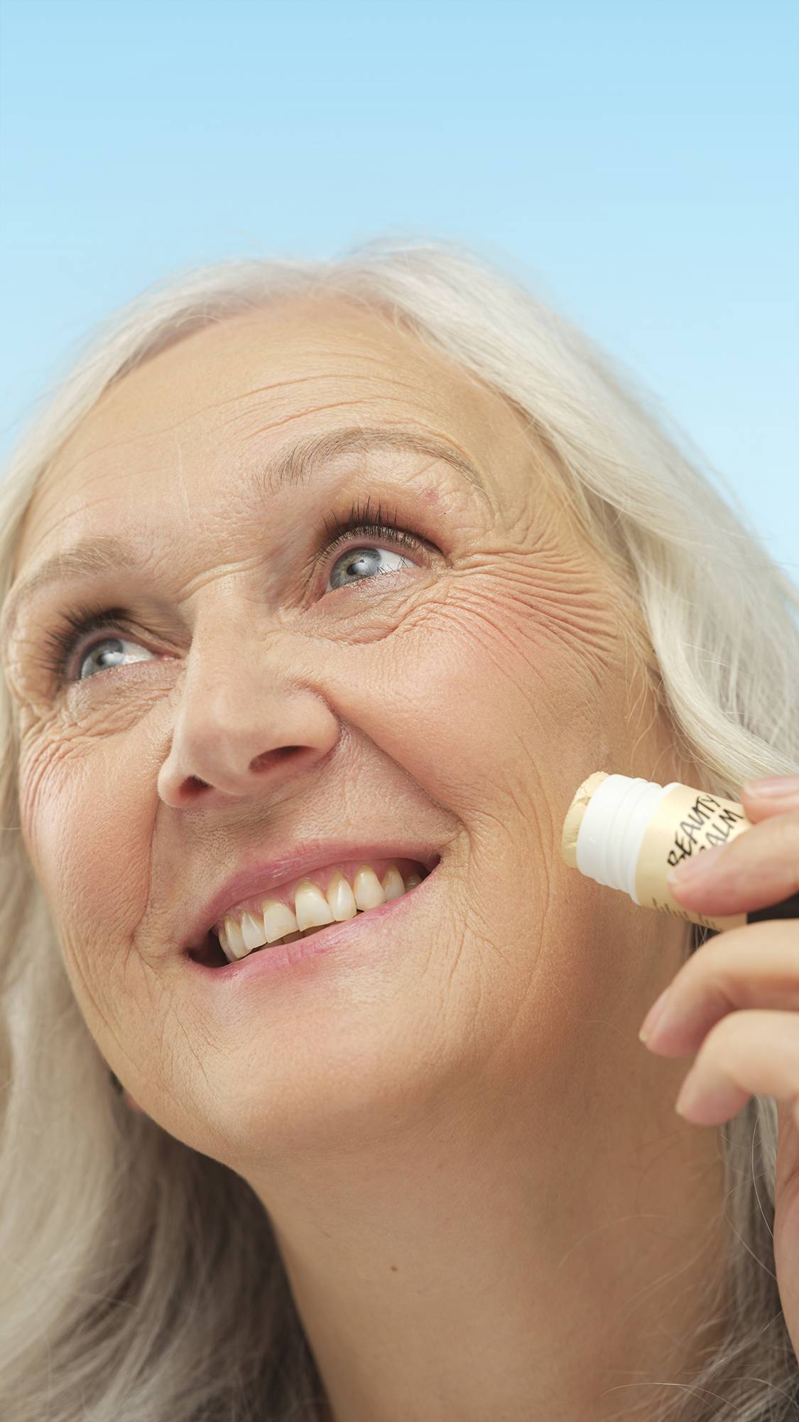 A close-up portrait of a smiling person with fair skin and white hair, looking up while applying the white stick of Light Neutral Beauty Balm directly to their cheek. The background is a bright blue.