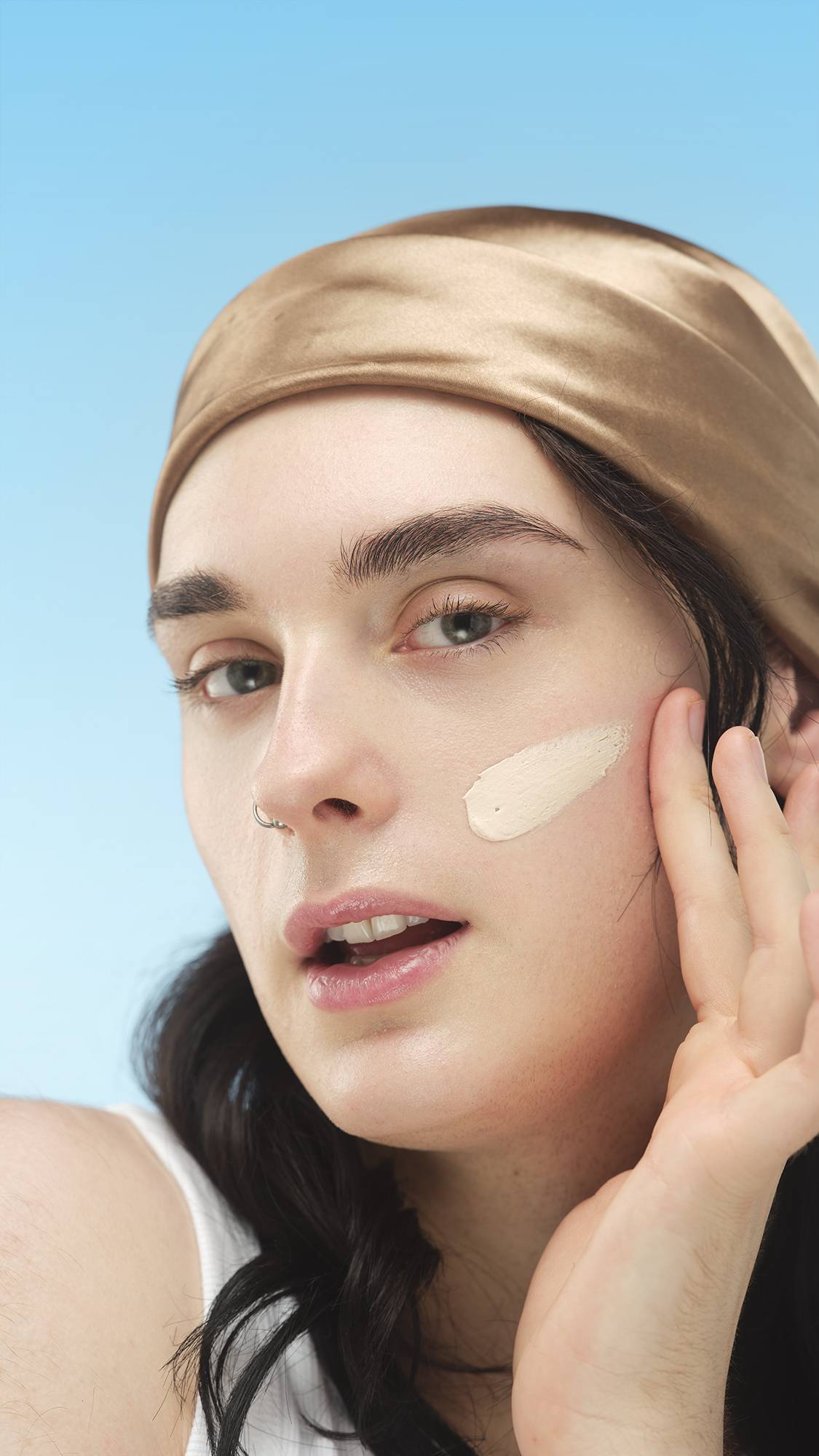 A close-up vertical portrait of a person with a golden-tan headband applying a swipe of Light Warm Skinwear cream to their cheek with their fingertip. The background is a solid light blue.