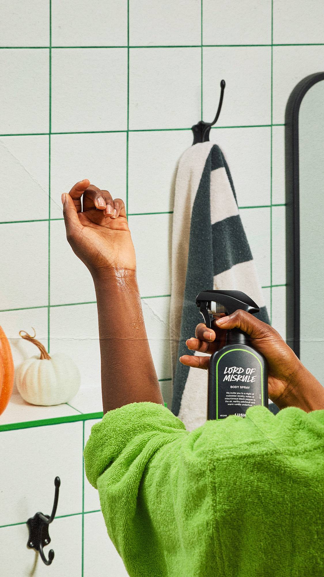 A model in a bright green dressing gown spritzes Lush Lord of Misrule body spray onto their raised forearm in a white-tiled bathroom with green grout, a striped towel, and small decorative pumpkins.