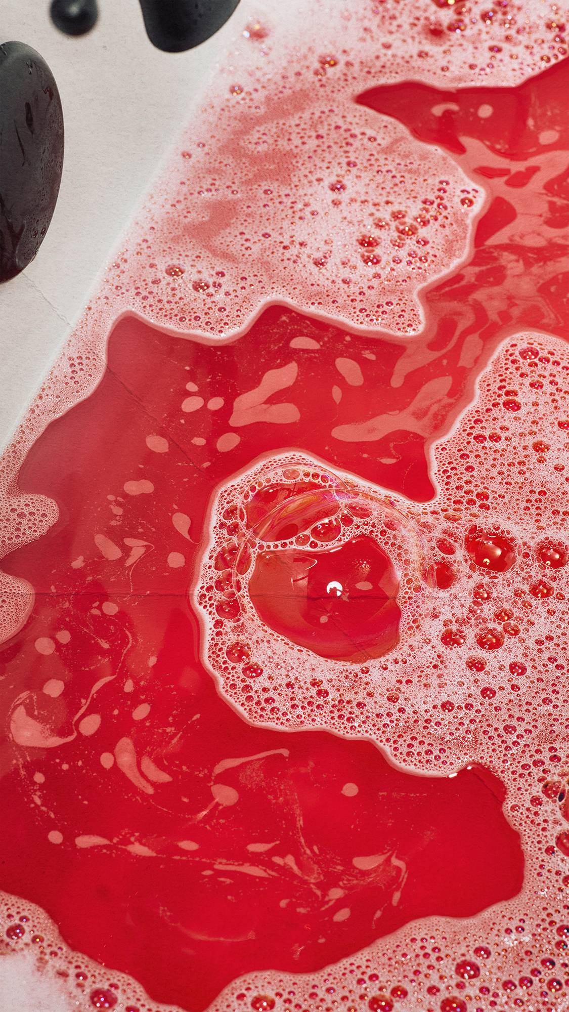 Close-up of vibrant, deep red bathwater covered in patches of fluffy white bubble bar foam, swirling together inside a white bathtub.