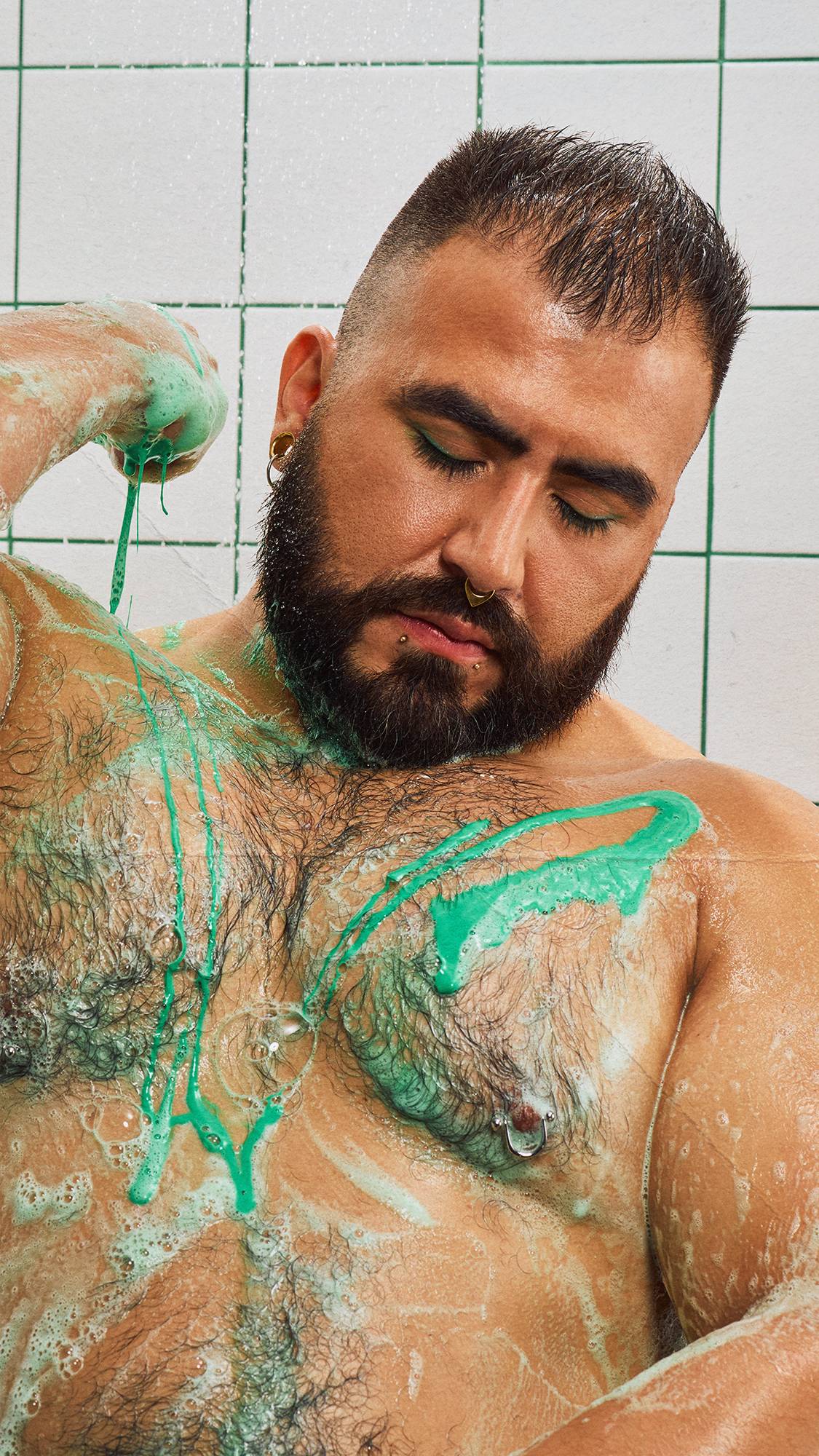 A bearded model with facial piercings stands with eyes closed in the shower, as bright green shower gel and soapy lather drip over their chest against a backdrop of white tiles with green grout.