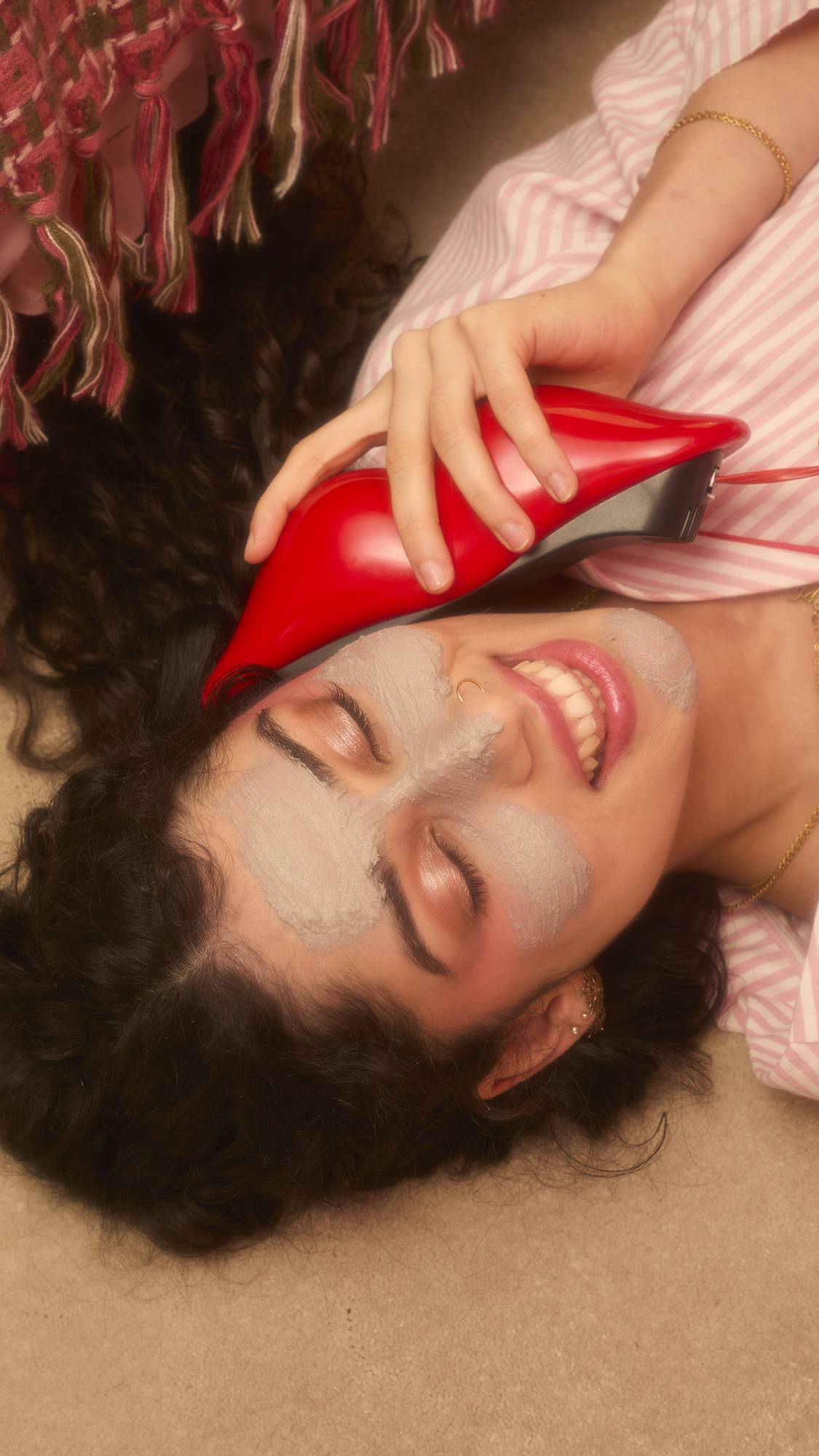 A smiling person lying on the floor in pink striped pajamas holds a red lip-shaped phone to their ear. Their face is covered in patches of the grey Lovey Dovey fresh facial cleanser.