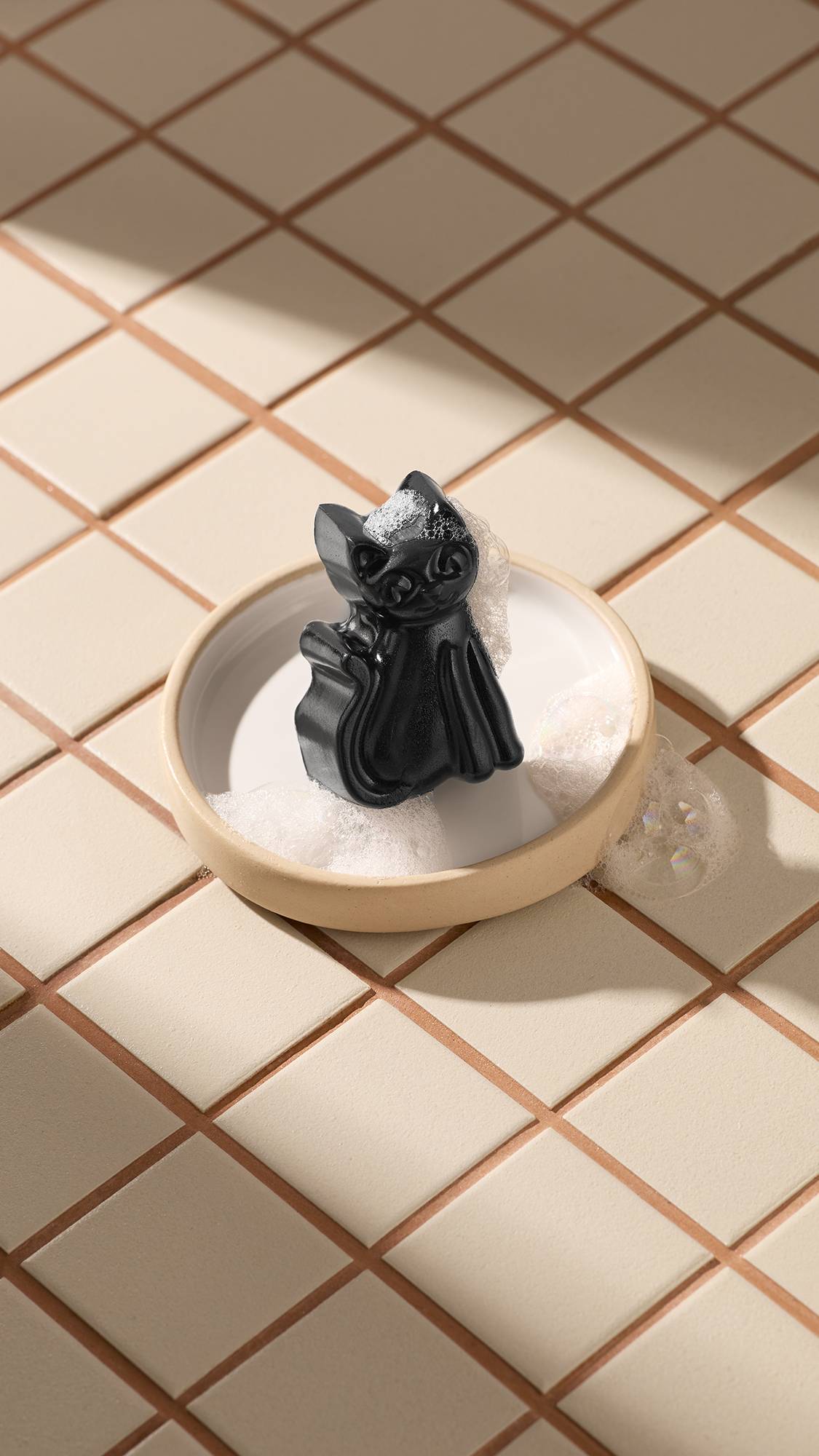 A black Lush Lucky Cat facial gummy sits in a round beige soap dish, surrounded by white soapy foam. The dish rests on light beige square tiles with brown grouting. Diagonal sunny shadows cross the scene.